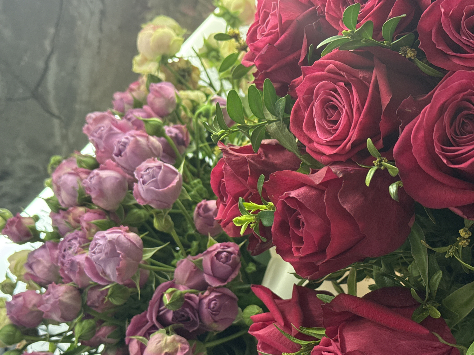 A close-up shot depicts an assortment of roses in varying colors - primarily a deep red and a soft lilac. Green foliage is interspersed amongst the blooms, suggesting a floral arrangement. The setting appears to be indoors. No specific location in Yerevan, Armenia can be deduced from the image. There are no people visible.