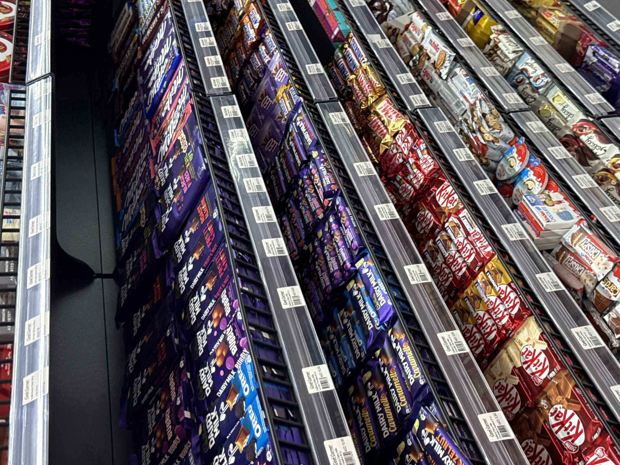 The image shows a close-up, high-angle view of a retail display filled with various chocolate bars and candy. The shelves are angled, creating a sense of depth and filling the frame. The primary subjects are the products themselves, with a significant variety of brands and packaging visible. Products include Dairy Milk bars in different flavors like Oreo, Bubbly, Fruit & Nut, and Caramel, along with Cadbury's Crunchie, Wispa, and Flake. Other prominent brands include Kit Kat bars in red and white wrappers, Snickers, and Kinder Surprise eggs.
The setting is clearly a supermarket or convenience store aisle, likely in Dubai, United Arab Emirates, given the price tags showing "AED 5.50" and "AED 2.50." The lighting appears to be artificial, common for indoor retail environments. There are no people visible in the image.
The activity depicted is the display of goods for sale. The arrangement suggests a well-stocked and organized retail space. The overall impression is one of abundance and variety of confectionery options. No specific time of day or weather conditions are discernible.
Visible text includes brand names such as "DAIRY MILK," "OREO," "BUBBLY," "FRUIT & NUT," "CARAMEL," "SNICKERS," "KIT KAT," and "KINDER SURPRISE." Price tags display "AED 5.50" and "AED 2.50." Some smaller text on the price tags appears to be in English and possibly Arabic, consistent with the location context.
