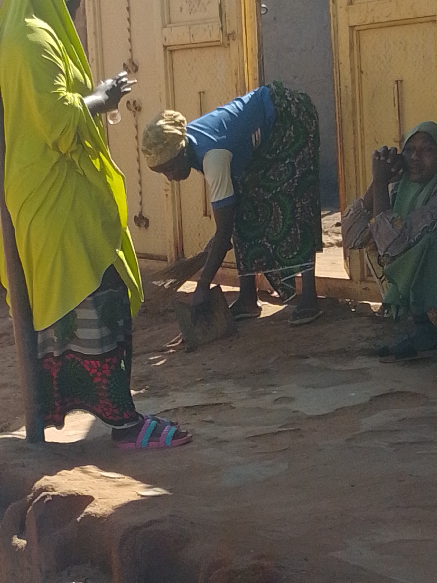 In an outdoor, unpaved area in Unknown, Unknown, three individuals are visible. On the left, a person draped in a bright yellow garment stands, holding a clear object in their right hand. To the right of this person, another individual wearing a blue shirt and a patterned skirt is sweeping the ground with a broom. Further to the right, a third person, wearing a green headscarf and a patterned garment, is seated and appears to be holding a mobile phone to their ear. The ground is sandy and uneven. In the background, there are two closed doors or gates, one yellow and one a lighter color, with decorative vertical elements. The scene is lit by natural sunlight, casting shadows on the ground.