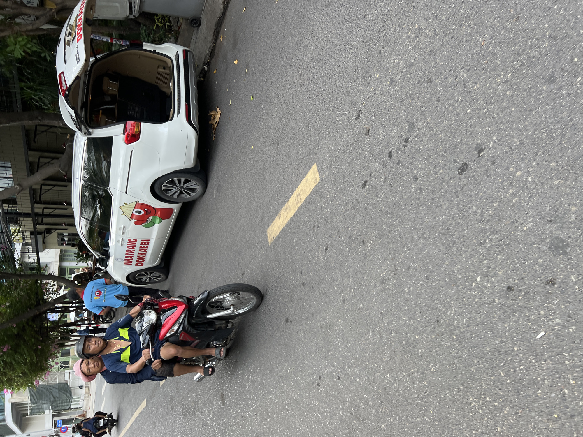 In Nha Trang, Vietnam, two individuals are traveling on a motorcycle on a city street. The rider, wearing a yellow and blue shirt, a helmet, and shorts, is driving. The passenger, a female wearing a pink head covering and a dark blue jacket, is seated behind the rider. They are on a red and black motorcycle.
Beside the motorcycle is a white minivan with its rear hatch open. The minivan has "NHATRANG DOKKAEBI" written in red on its side, along with a cartoon character. A man in a blue shirt with a logo is standing near the minivan. The scene takes place on an asphalt street with a yellow line marking. Trees line the street, and buildings are visible in the background. The overall environment appears to be an urban setting.