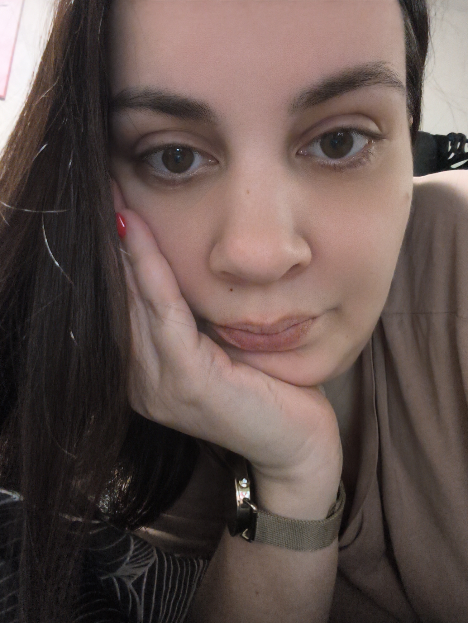 An adult female is depicted in a close-up, head-and-shoulders portrait, gazing directly into the camera. She possesses dark brown hair, some strands of which feature light-colored highlights, flowing over her left shoulder and face. Her eyes are brown, and her eyebrows are dark. A small dark mark is visible on the bridge of her nose. Her lips are a light reddish-brown, slightly parted in a neutral expression. Her left hand is raised, with fingers supporting her chin and jawline. The fingernails on her visible fingers are painted red. She is wearing a light tan or beige-colored top, exhibiting visible fabric folds. On her right wrist, a gold-toned watch with a metallic mesh band is present. The background is indistinct and out of focus, suggesting an indoor setting, with faint lighter and darker areas visible, including a dark object with white linear patterns on the right. The scene is located in the city of Chaikovsky, Russia.