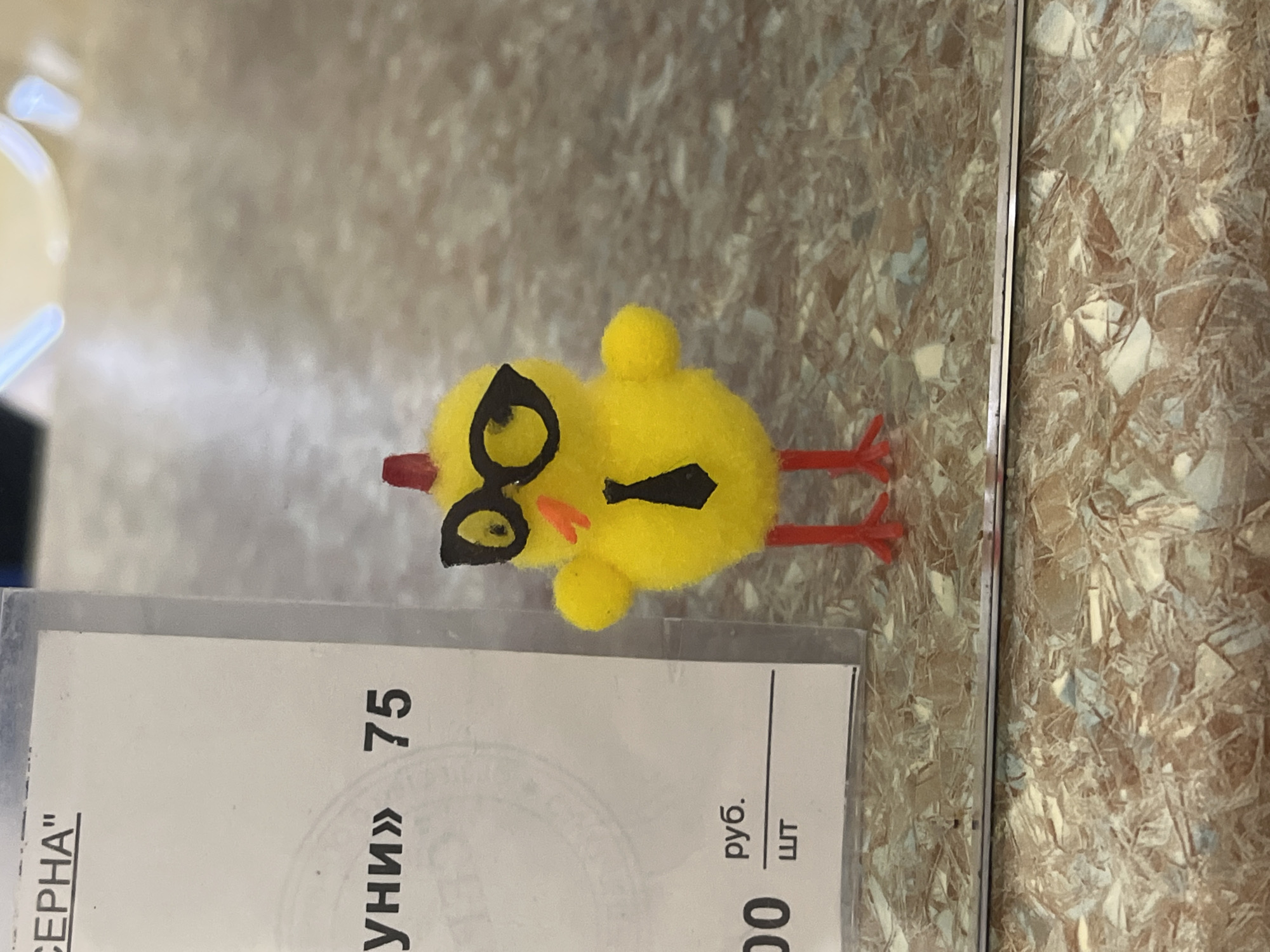 This image captures an indoor scene, rotated 90 degrees clockwise, featuring a small, bright yellow chick toy resting on a textured surface. The chick, made of fuzzy material, is adorned with small black spectacles, a tiny black tie, and has a red comb-like tuft on its head and two thin red stick-like legs. It appears to be lying on its side on the surface.
Adjacent to the chick is a piece of white paper, partially visible and covered by a clear plastic protective sleeve. The paper displays text in Cyrillic script, oriented sideways due to the image rotation. Visible text includes "СЕРНА", possibly part of a product name, and "УНИ» 75". Further down, the Russian currency abbreviation "руб." (ruble) and "ШТ" (shtuka, meaning 'piece' or 'item') are discernable, along with the digits "00", likely indicating a price or quantity. These Russian details are consistent with the Saint Petersburg, Russia context.
The background surface is light brown with a complex, granular texture resembling compressed wood chips or a laminate pattern mimicking it. There are no people visible, and no overt activity is taking place; it is a static composition of objects. The lighting suggests an indoor setting, possibly under artificial light.