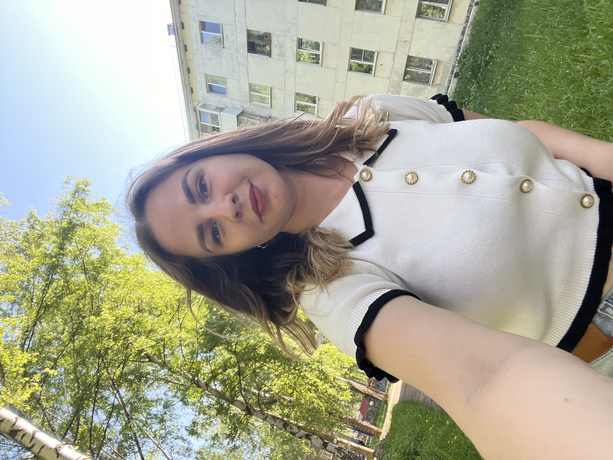 A selfie shows a woman in Izhevsk, Russia, with a pale complexion, light brown hair, and light eye color. She is wearing a white, knit, short-sleeved top with black trim and pearl buttons. Behind her, a multi-story, light-colored building with evenly spaced windows is visible. To the left, there are birch trees with green foliage. The sky is visible in the upper-left corner and is a clear, light blue. Green grass covers the ground throughout the background.