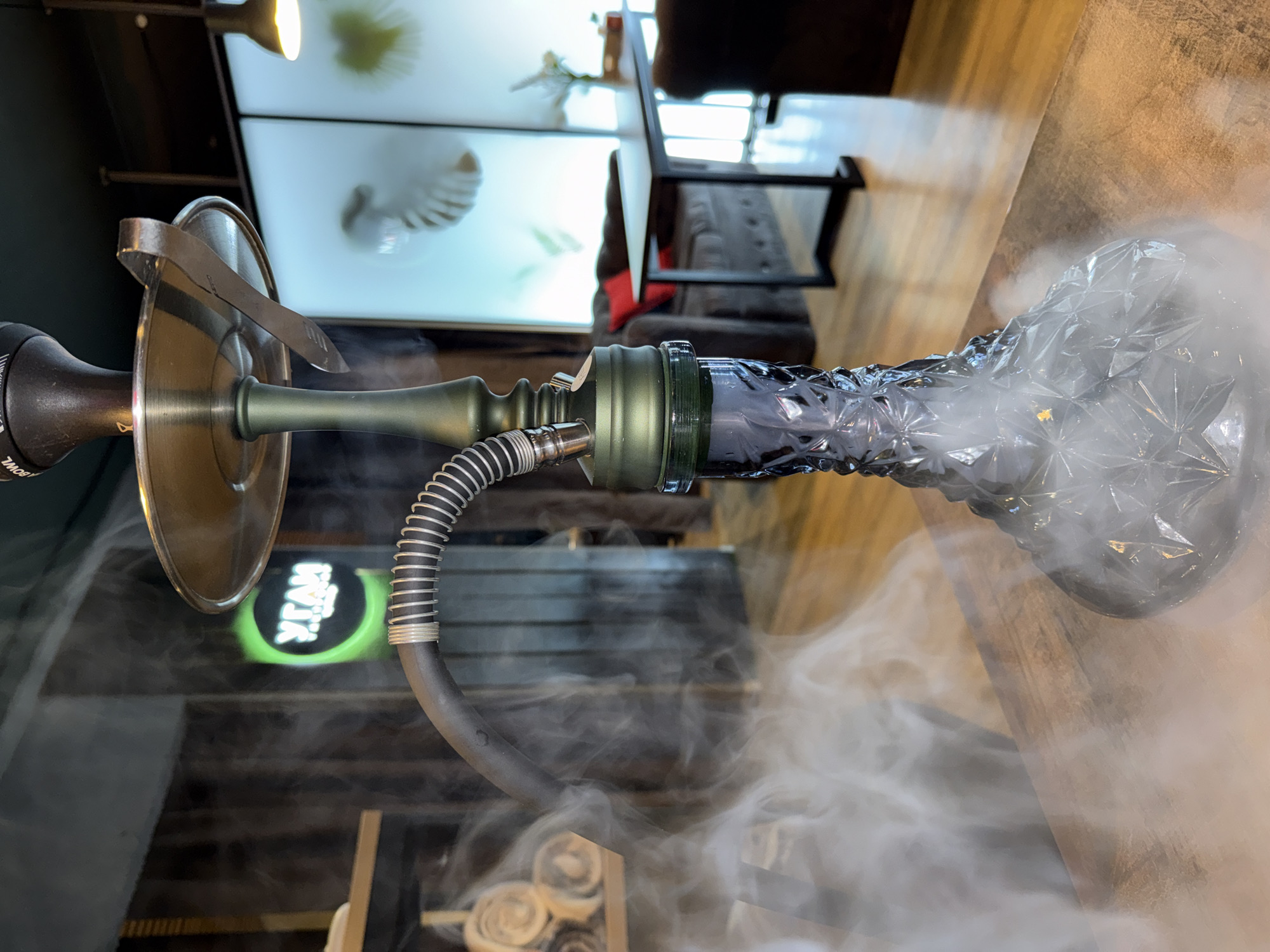 An indoor scene features a prominent hookah resting horizontally on a surface, likely a table. The hookah has an olive-green metallic stem and a large, round, metallic tray with charcoal tongs resting on it. A black, ribbed hose with a silver coiled section connects to the stem. The hookah's base is made of clear, faceted glass, resembling a diamond cut pattern, and is filled with a dense white smoke that also billows around the base and hose, indicating active use.
In the background, out of focus, are elements of a room, possibly a lounge or cafe. A dark sofa or armchair is visible, along with warm-toned wooden surfaces. On a wall, two large, brightly lit rectangular panels display blurry, light-colored, abstract shapes. An overhead light fixture casts illumination from the top left.
A notable detail is a partially visible green circular logo with white Cyrillic text, which appears to read "УГЛИ" (meaning "coals" in Russian), reinforcing the location context of Chaikovsky, Russia, and suggesting the setting is a hookah lounge. No people are visible, but the presence of abundant smoke implies recent or ongoing activity. The image has a warm, ambient lighting, typical of an evening setting.