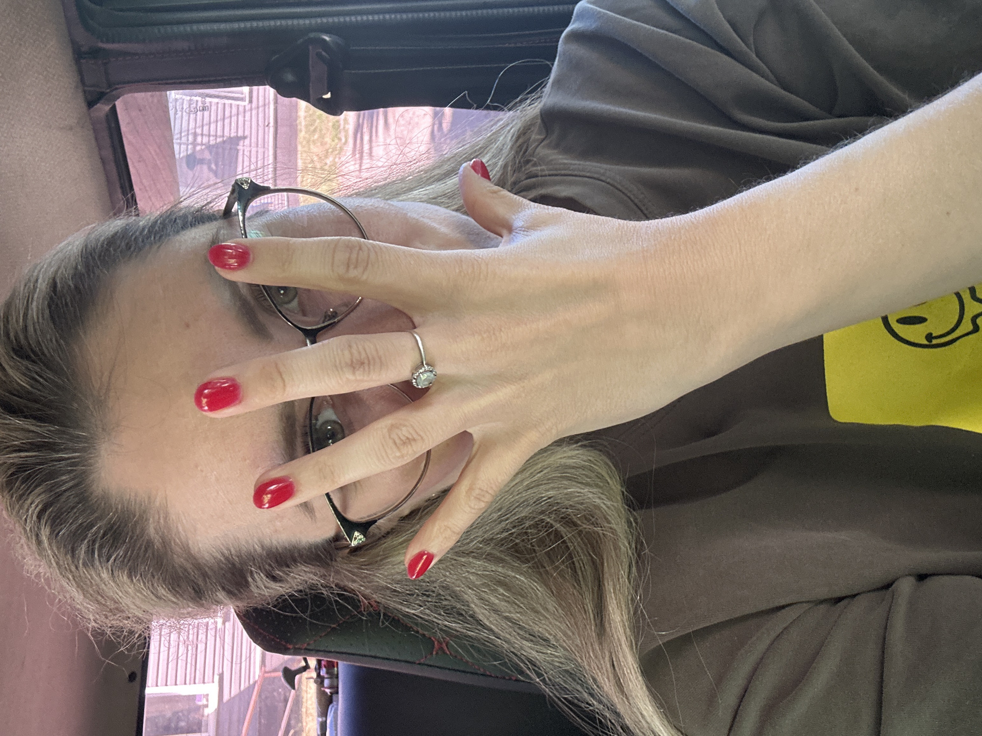 A woman is captured in a close-up shot from the chest up, seated inside a vehicle. Her right hand is raised, obscuring the upper portion of her face. She has long, blonde hair and is wearing glasses. Her fingernails are painted bright red, and a ring with a single clear stone is visible on her left ring finger. She is wearing a brown t-shirt with a yellow graphic on the left side. The background suggests an outdoor setting with pink-tinted windows and a building visible through them, indicating a location likely in a city or town. The specific location of Novoaltaysk, Russia, is not identifiable from the visual information present in the image.