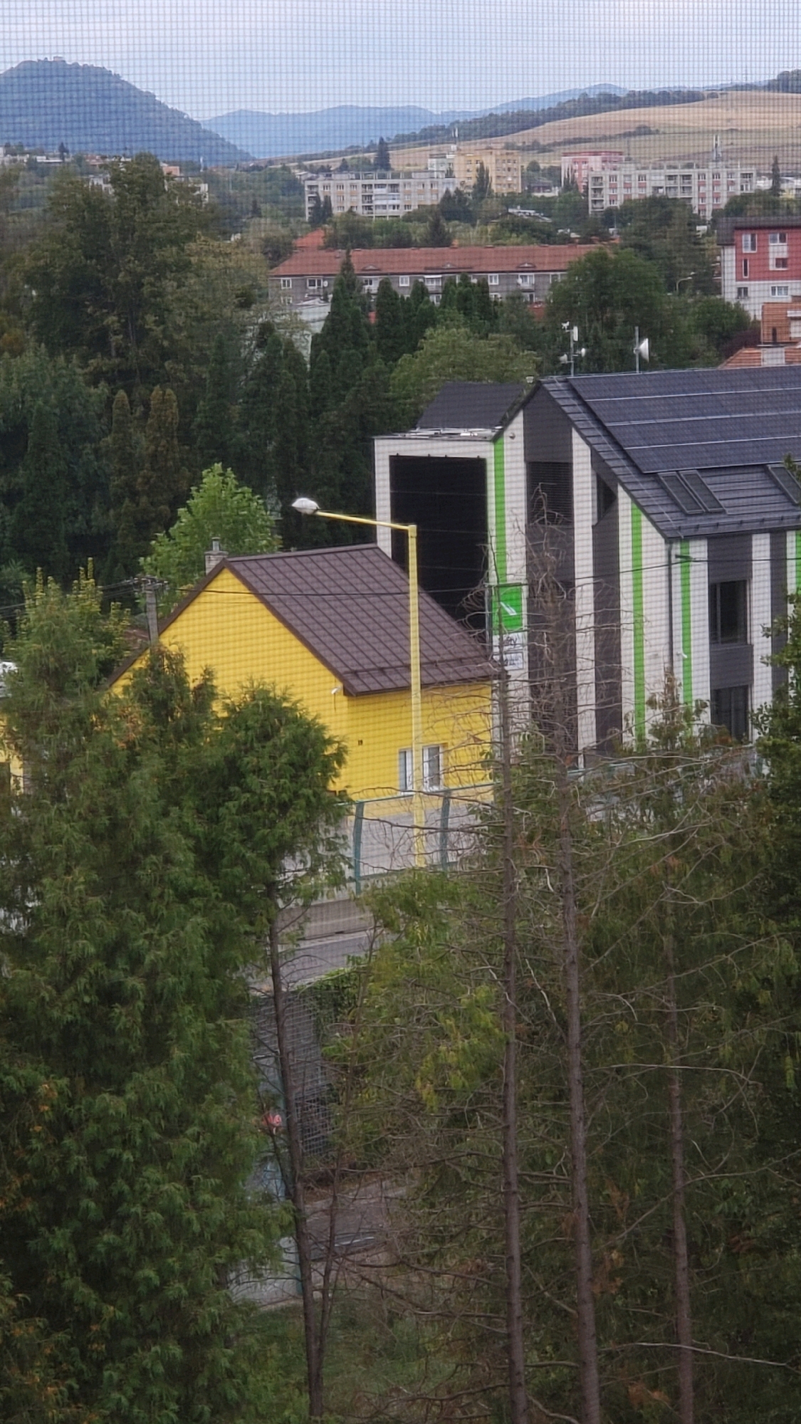 This is a medium-distance, eye-level, outdoor, daytime photograph taken from an elevated position looking down and across a residential area in Prešov, Slovakia. The foreground is dominated by a dense cluster of evergreen trees, with some deciduous trees interspersed. Beyond the trees, a bright yellow, single-story house with a dark brown roof is visible. To the right of the yellow house, a modern, multi-story building with a dark roof featuring solar panels and a facade of white, black, and green vertical stripes is partially visible. A yellow street lamp stands in front of the yellow house. In the background, rolling hills and mountains are visible, with a cityscape of apartment buildings and other structures situated between the foreground and the hills. The sky is overcast. The image appears to be taken through a fine mesh, possibly a window screen or netting. No people or animals are clearly visible.