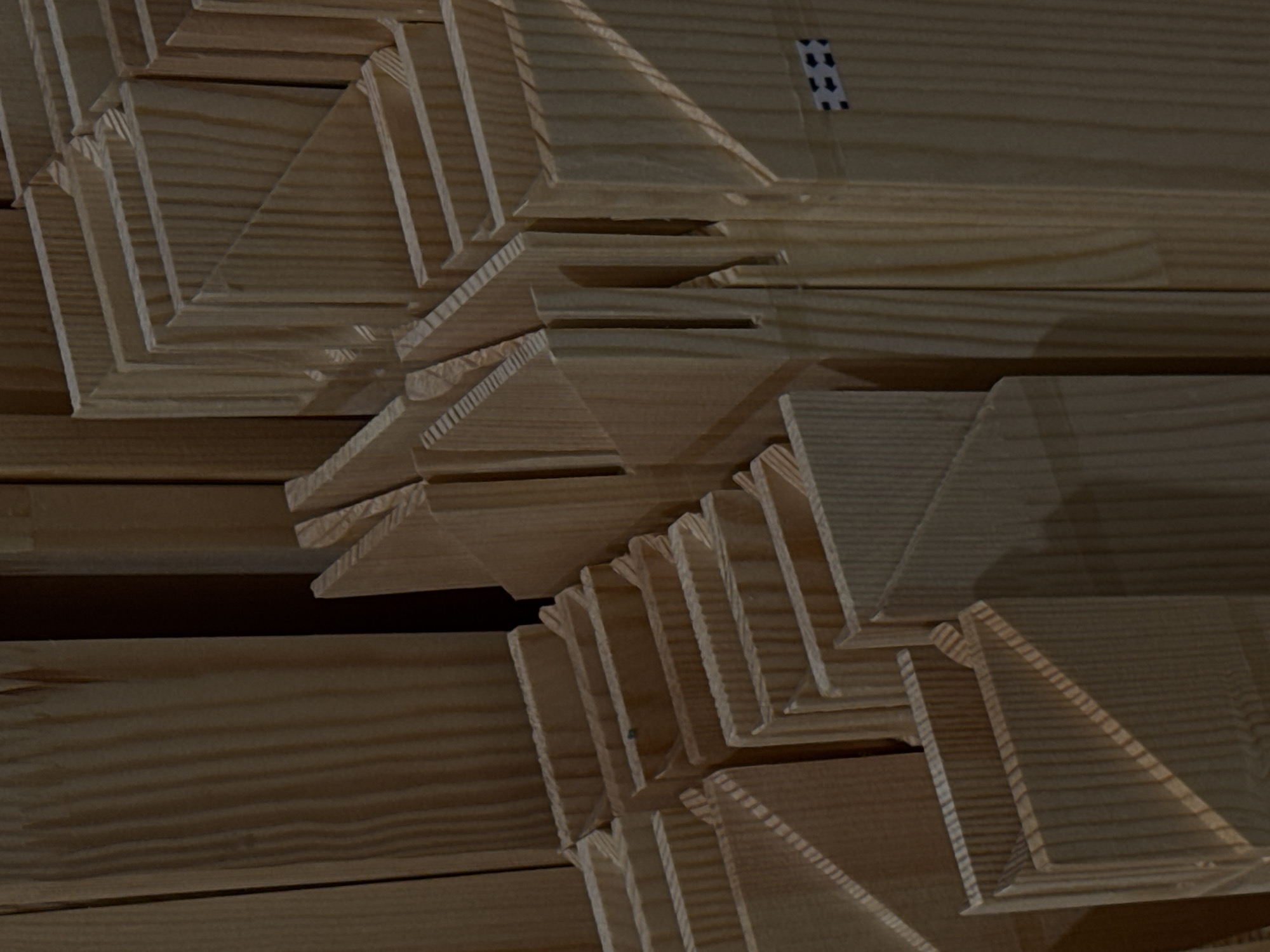 The image is a close-up, dimly lit shot of stacked wooden planks, likely lumber, in a workshop or storage area. The wood grain is prominent, and the planks appear to be unfinished, with visible cuts and grooves. Some planks are arranged in a way that suggests they might be components for a larger structure, perhaps frames or shelving. There are no people visible in the image. The lighting creates strong shadows, highlighting the texture of the wood. The scene is devoid of any identifiable landmarks or features that would indicate its specific location in Moscow, Russia, or anywhere else. The image is entirely focused on the texture and arrangement of the wooden materials.