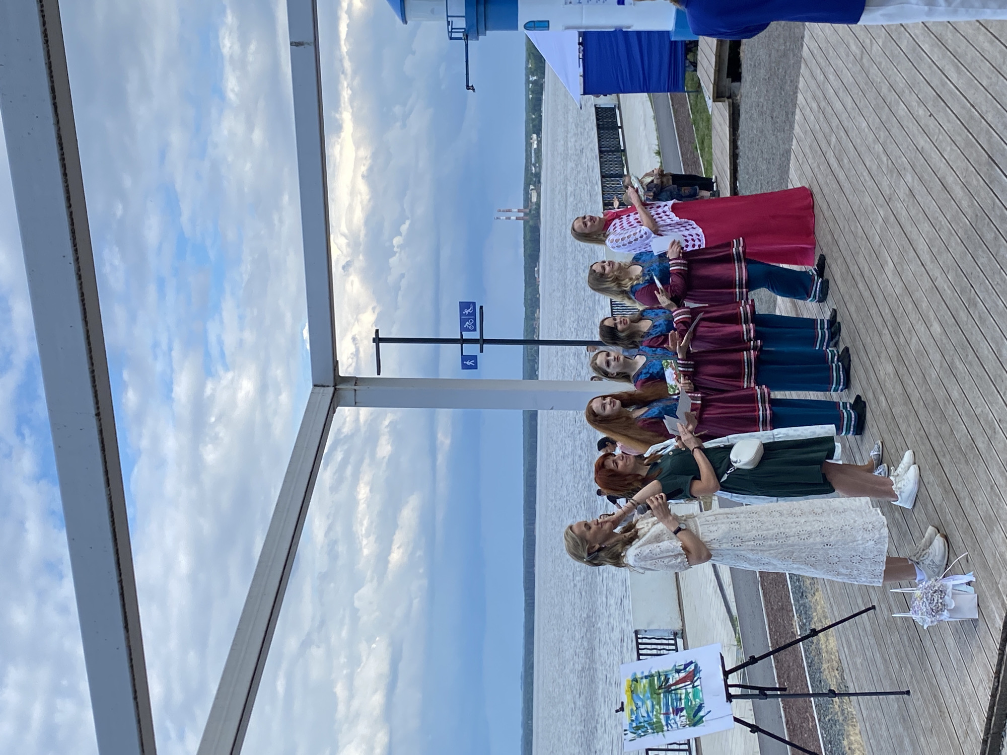 The image shows a group of people in Chaikovsky, Russia, on what appears to be a wooden boardwalk next to a body of water. The sky is cloudy, suggesting it could be daytime with overcast weather.
The main subjects are a line of women, some of whom are wearing traditional-looking outfits with dark red and blue elements. One woman in the foreground is wearing a white lace dress and appears to be speaking into a microphone. Next to her is an easel displaying a painting. Another woman in a dark green dress is also holding a microphone. The other women in line are dressed in matching burgundy and teal garments, some holding papers or programs.
The setting is outdoors, near a large body of water, possibly a river or lake. In the background, there are structures that resemble a lighthouse or observation tower, and further in the distance, industrial chimneys. The overall mood seems to be that of an outdoor performance or cultural event. The lighting indicates it is likely daytime. There are no visible signs with text.