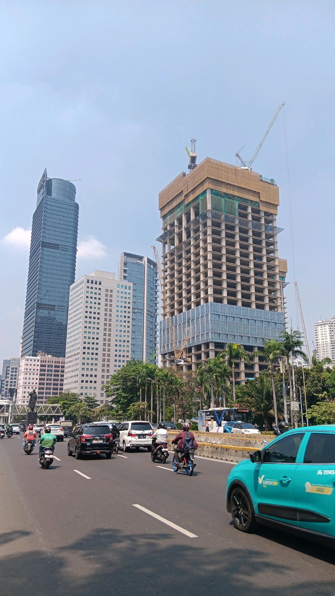 In this mid-angle, eye-level shot, a bustling city scene in Central Jakarta, Indonesia, unfolds under a bright, partially cloudy sky. The foreground features a multi-lane asphalt road with several vehicles. A turquoise car, identified by the "GREEN.SM" logo and license plate "JG 11145", occupies the right side of the frame. To its left, a motorcyclist wearing a helmet and dark jacket rides a blue motorcycle, followed by another motorcyclist on a black motorcycle. Further down the road, a black SUV and a white MPV are visible, along with more motorcyclists.
The background is dominated by a construction site, with a tall building in the process of being erected. Two large construction cranes are positioned around the unfinished structure, one reaching high into the sky. Adjacent to the construction site stands a very tall, dark blue glass skyscraper. To its left, a lighter colored, multi-story building with numerous windows is also prominent, followed by a shorter, modern office building. Green trees and palm trees line the median and the sides of the road, interspersed with the urban architecture. A pedestrian bridge can be seen in the distance, and a distant white building suggests further development in the cityscape. A statue on a pedestal is also visible on the left side of the image, near the tall blue skyscraper.