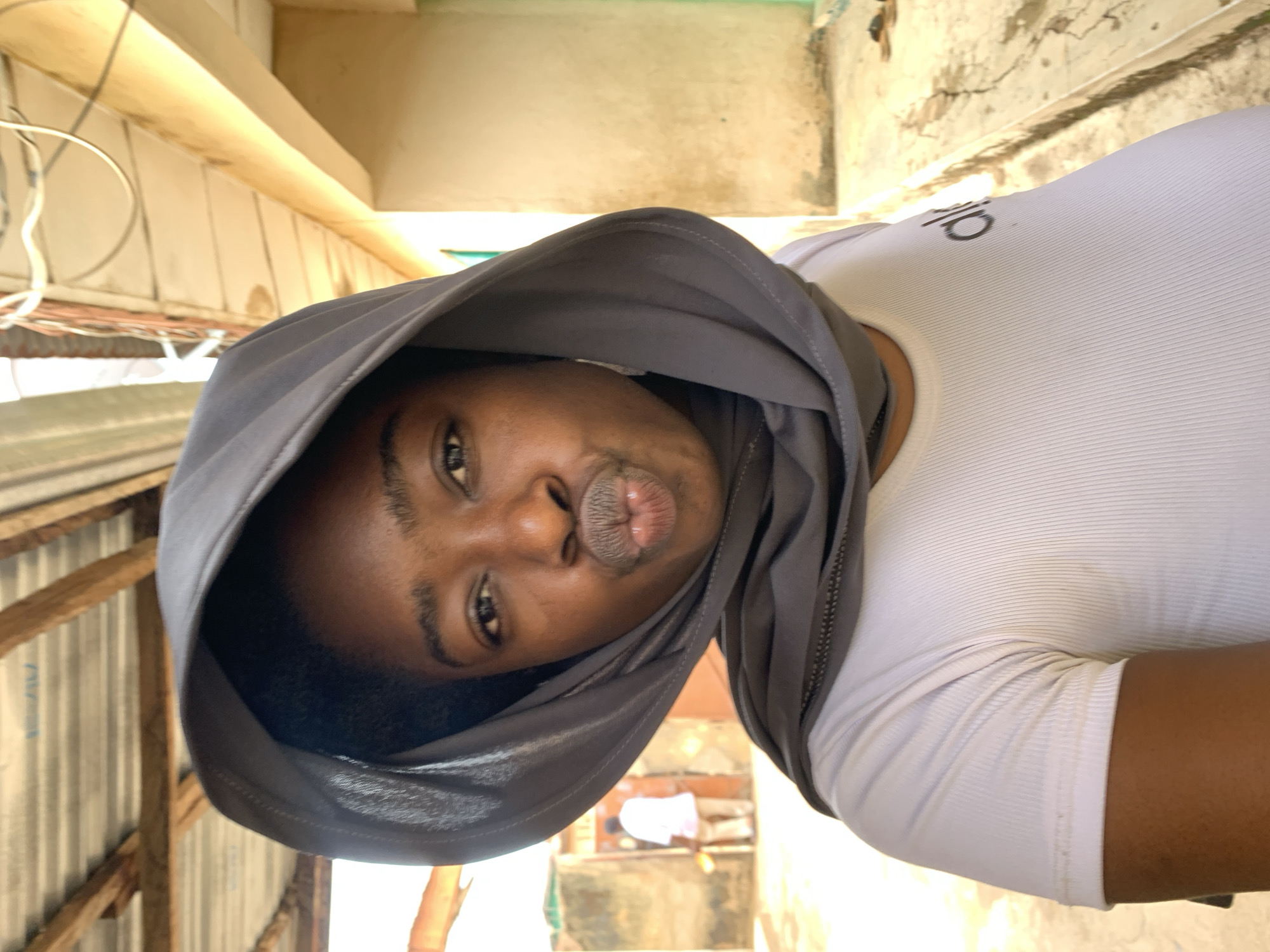 A woman with dark skin, wearing a grey head covering and a white ribbed shirt with a logo visible on the upper left, is posing for a photo. Her lips are pursed in a kiss. The environment suggests an outdoor setting in a residential area, possibly in Rafin Par, Nigeria. Corrugated metal and wooden beams are visible to the left, suggesting a makeshift or informal structure. In the background, there are glimpses of other buildings and what appears to be clothing hanging to dry.
