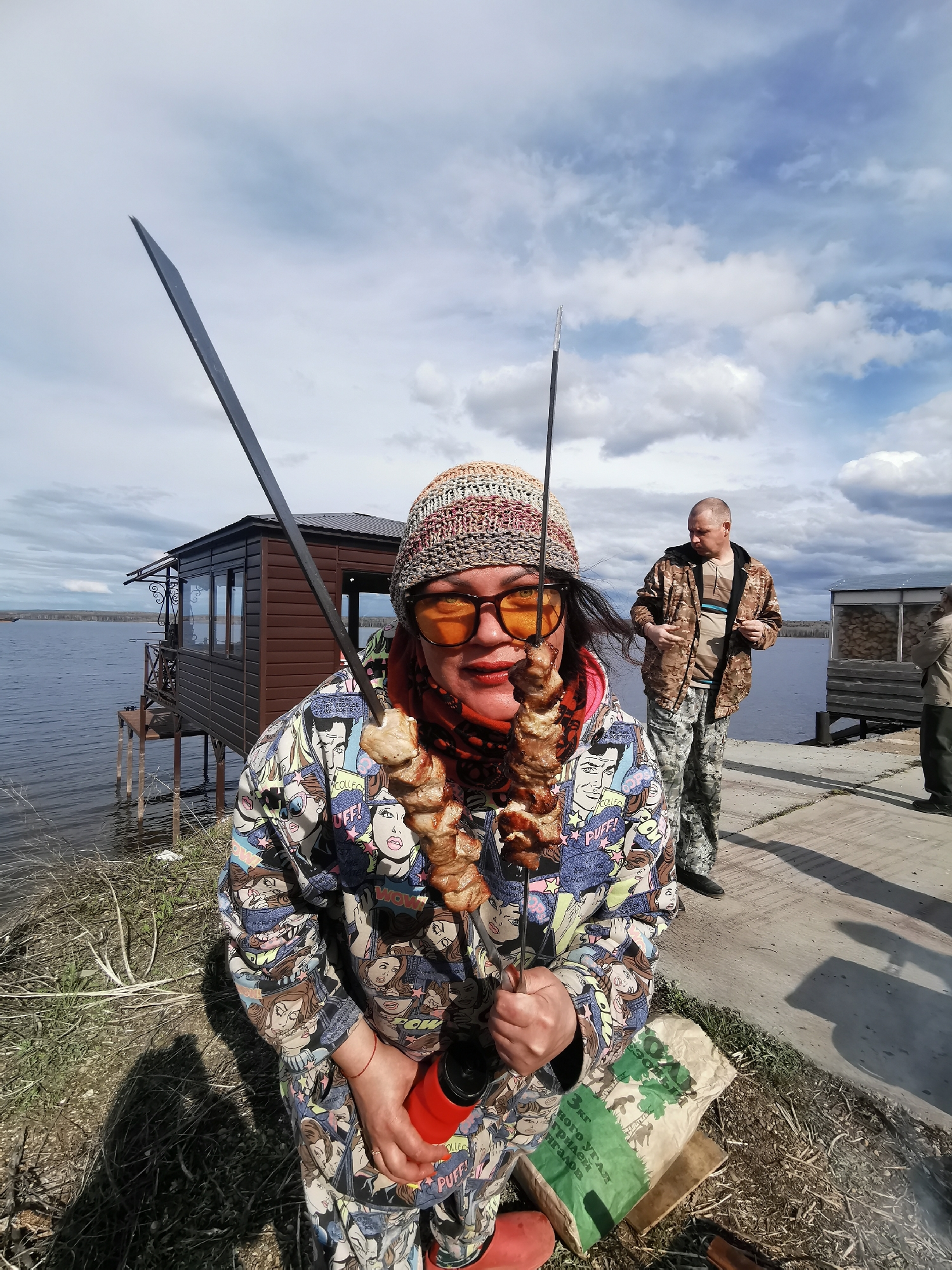 A daytime outdoor scene captures a woman holding skewers of meat, smiling at the camera. She wears a colorful knitted beanie, orange-tinted sunglasses, and a vibrant jacket adorned with pop art-style comic strip faces. A red scarf is visible beneath her jacket. In her left hand, she holds a red water bottle, while her right hand holds two metal skewers. One skewer, held lower, is laden with uncooked meat cubes, likely for shashlik or kebab. The other, held higher and angled upwards, appears to be an empty skewer or perhaps another type of grilling tool.
Behind the woman, a man in camouflage jacket and pants stands on a concrete pier, looking to his right. Further in the background, another man is partially visible. The setting is by a large body of water, possibly a lake or wide river, with a small brown wooden building on stilts extending over the water on the left side of the frame. The sky above is overcast with patches of blue and white clouds.
At the bottom right, a green paper bag of charcoal rests on the ground. Visible text on the bag in Cyrillic reads "3кг ДРЕВЕСНОГО УГЛЯ для гриля и шашлыков" (3kg WOOD CHARCOAL for grill and shashliks), confirming the grilling activity. The ground around the woman is a mix of dirt and dry grass. Shadows are visible on the ground, indicating daylight. The overall atmosphere suggests a casual gathering or picnic, centered around outdoor cooking.