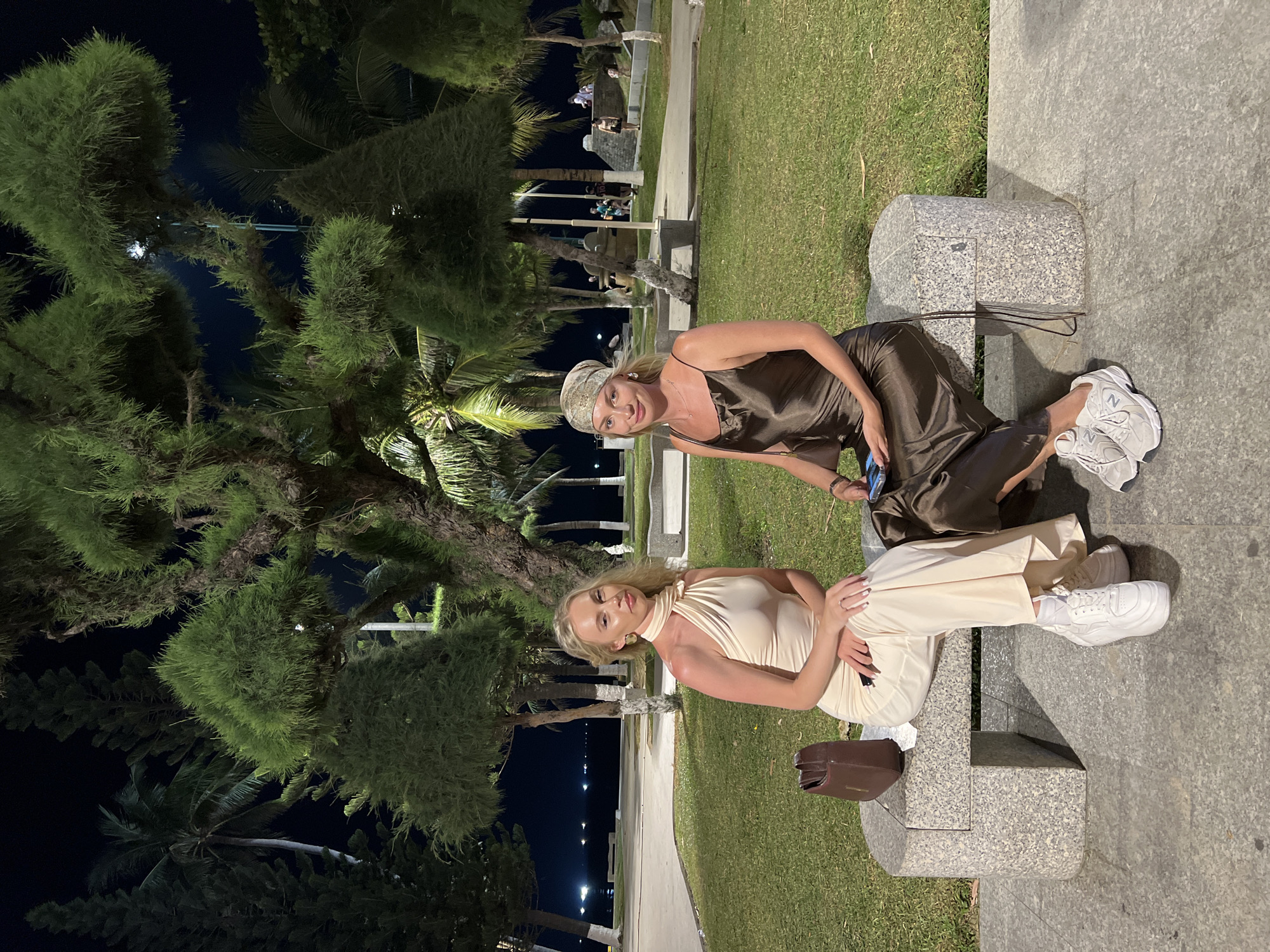 Two women are sitting on a stone bench in Nha Trang Ward, Vietnam. One woman is wearing a satin dress and a headscarf, and the other woman is wearing a halter-neck dress and pants. They are both wearing sneakers. A brown purse is on the bench next to the woman in the light dress. The bench is surrounded by grass. Behind them, there are palm trees and street lights.