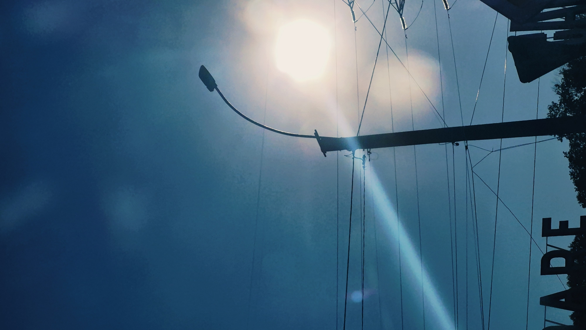 The image shows a silhouette of a lamppost against a bright sky. The sun is visible as a bright orb in the upper right quadrant of the image, casting a lens flare across the sky. Several power lines crisscross the frame in front of the lamppost and the sky. In the lower left corner, the silhouette of large letters spelling "VAPE" is visible, suggesting the image may have been taken in front of a vape shop. The presence of trees at the bottom edge of the frame indicates an outdoor setting. The overall impression is one of a street scene, possibly in an urban environment. Without additional information or context, it is impossible to determine the specific location within Chișinău, Moldova.