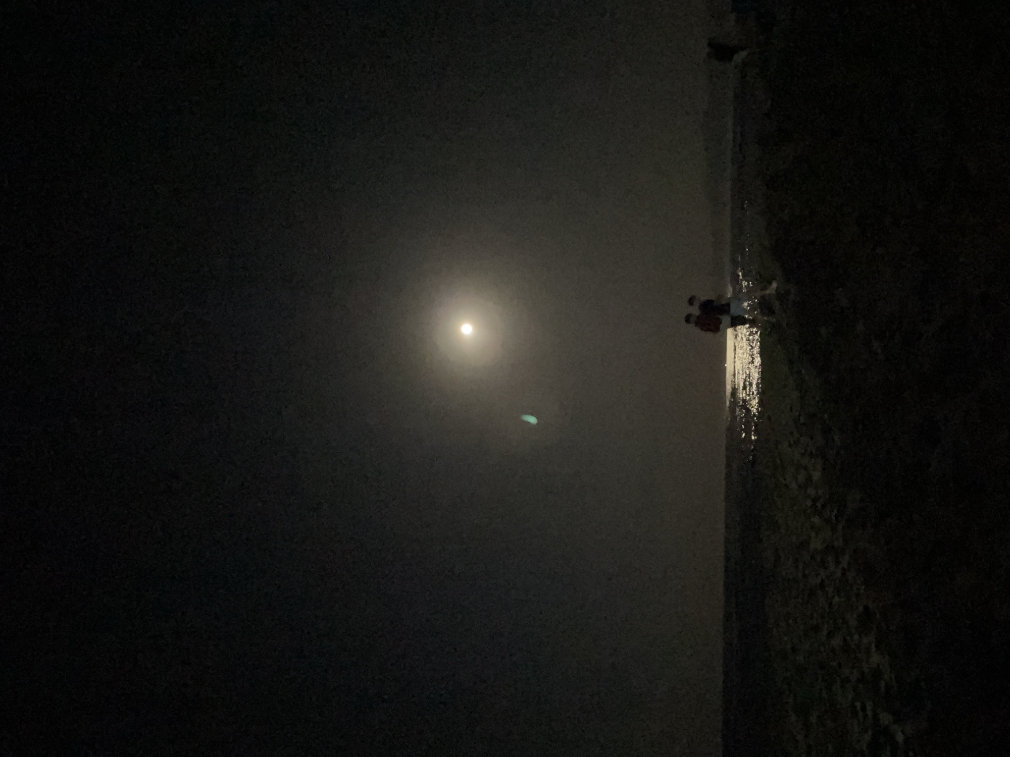 At night, under a full moon, two individuals stand on a beach. The moon is the brightest object in the sky, casting a bright reflection on the water. The beach appears to be made of sand. One individual is wearing a red jacket, and the other is wearing a dark shirt and white shorts. There is another person in the background, further to the right. The overall atmosphere of the image is dark and serene. The location is not identifiable as being in the city of Unknown, Greece.