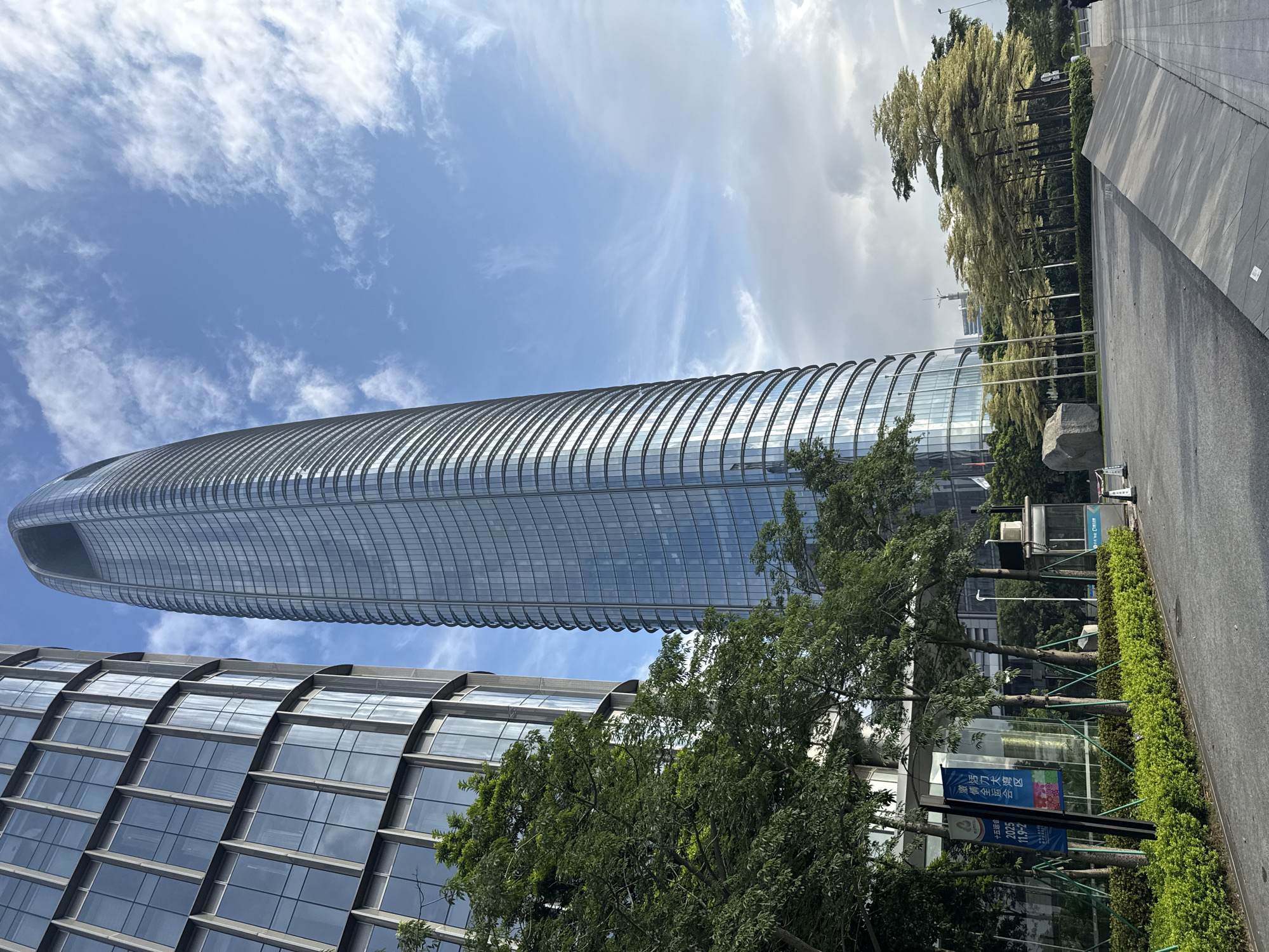 The image captures a street-level view of a modern urban environment in Haizhu District, China, likely during the daytime given the bright sky and shadows. Dominating the scene are two contemporary skyscrapers with sleek, glass facades. The taller building on the right is distinctively curved, tapering towards its apex, with a series of horizontal bands of windows and reflective surfaces. To its left, another building displays a more rectilinear, though also curved, facade with a grid of rectangular windows.
In the foreground, a paved walkway leads towards the buildings, bordered by a neatly manicured hedge and several young trees with lush green foliage. Beyond the hedge, a series of taller trees with a distinctive canopy shape, some appearing to have yellow-green leaves, are arranged in a line. A large, grey boulder rests on a grassy area in front of the curved skyscraper. Several flagpoles stand nearby.
A blue sign with Chinese characters is visible on the left, accompanied by smaller informational signs. A small, enclosed booth, possibly a security or information kiosk, is positioned on the right side of the walkway. The sky above is a vibrant blue, dotted with white, wispy clouds, suggesting fair weather. The overall impression is one of a well-maintained, modern commercial or civic area.