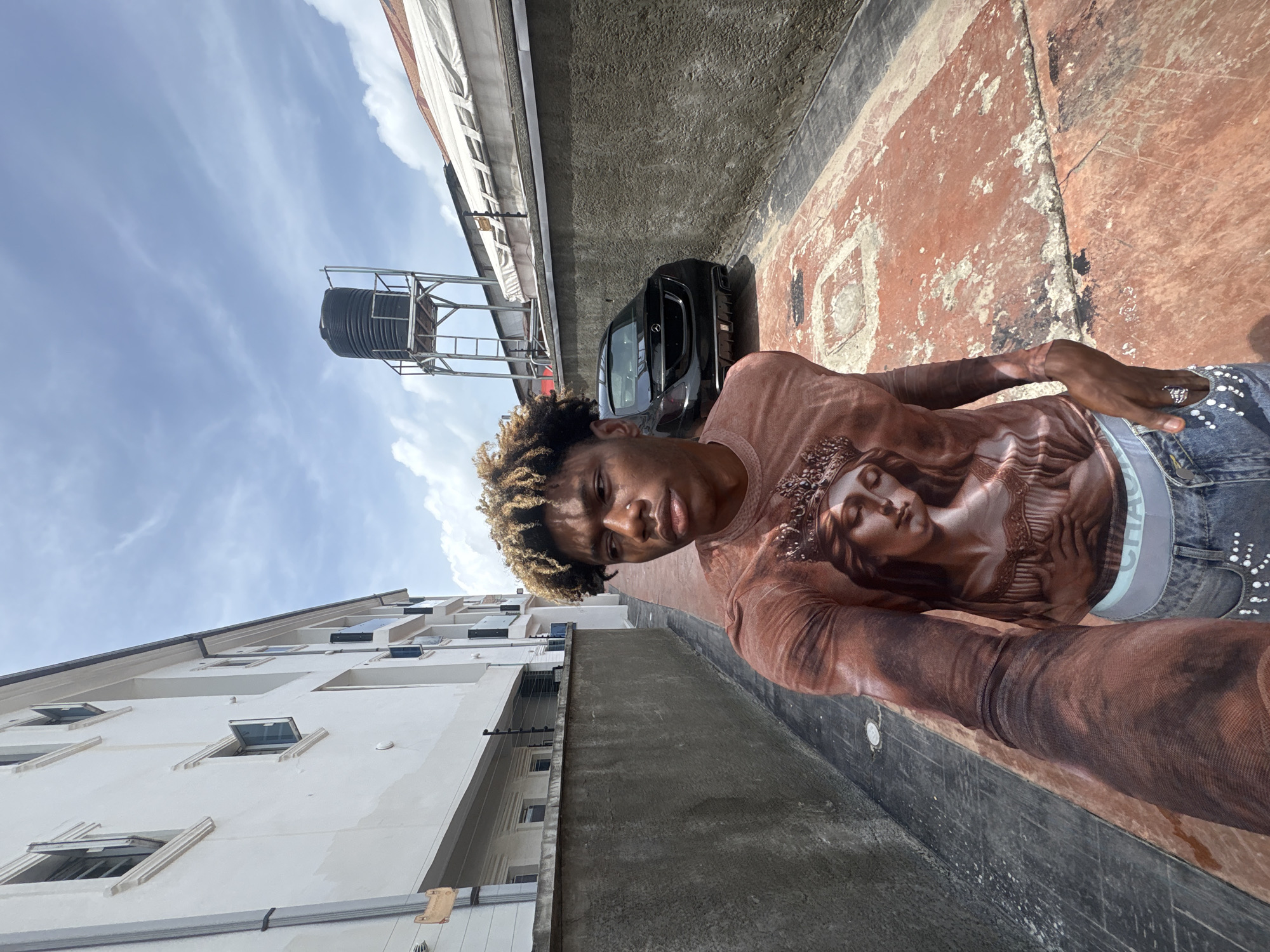A young man stands on a rooftop in Igboefon, Nigeria, taking a selfie. He is wearing a brown, patterned top and jeans. Behind him, there is a multi-story white building with windows, a black car, a water tower, and a reddish-brown rooftop surface. The sky is visible in the background, with scattered clouds.