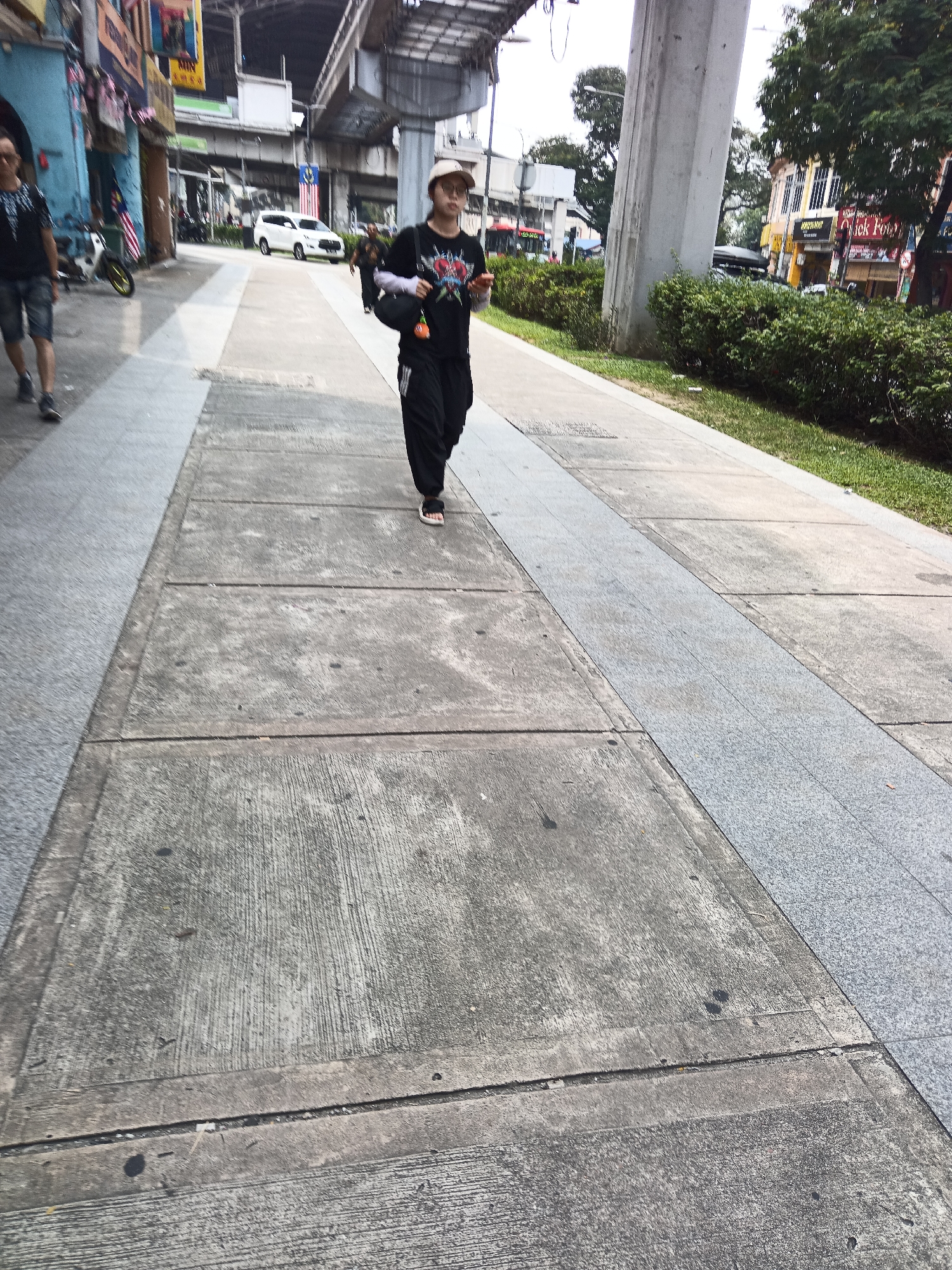 The image captures a pedestrian sidewalk in Kuala Lumpur, Malaysia, beneath an elevated transit line. A person wearing a beige cap, glasses, a black t-shirt with a graphic print, and black pants is walking towards the camera. They are carrying a black bag on their left shoulder and holding a light-colored object in their right hand. To the left of the frame, another person is visible walking away from the camera, wearing a dark t-shirt and shorts. The sidewalk is paved with concrete slabs and features a decorative granite strip. In the background, there are buildings with colorful facades, parked cars, and lush green bushes lining the street. The overall scene suggests a bustling urban environment.