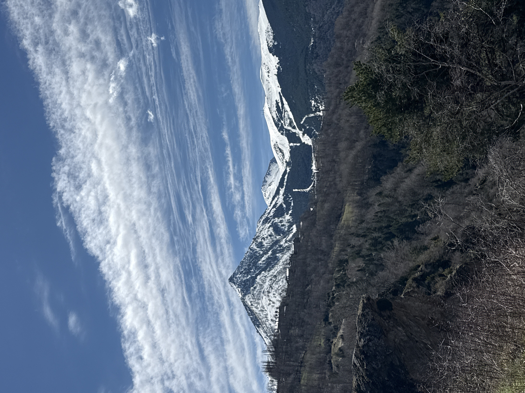 The image shows a mountain landscape near Benasque, Spain. The sky is blue with streaky white clouds. A steep mountain covered with snow in patches rises on the left, transitioning into a forested slope to the right. The foreground consists of rock formations and bare trees.