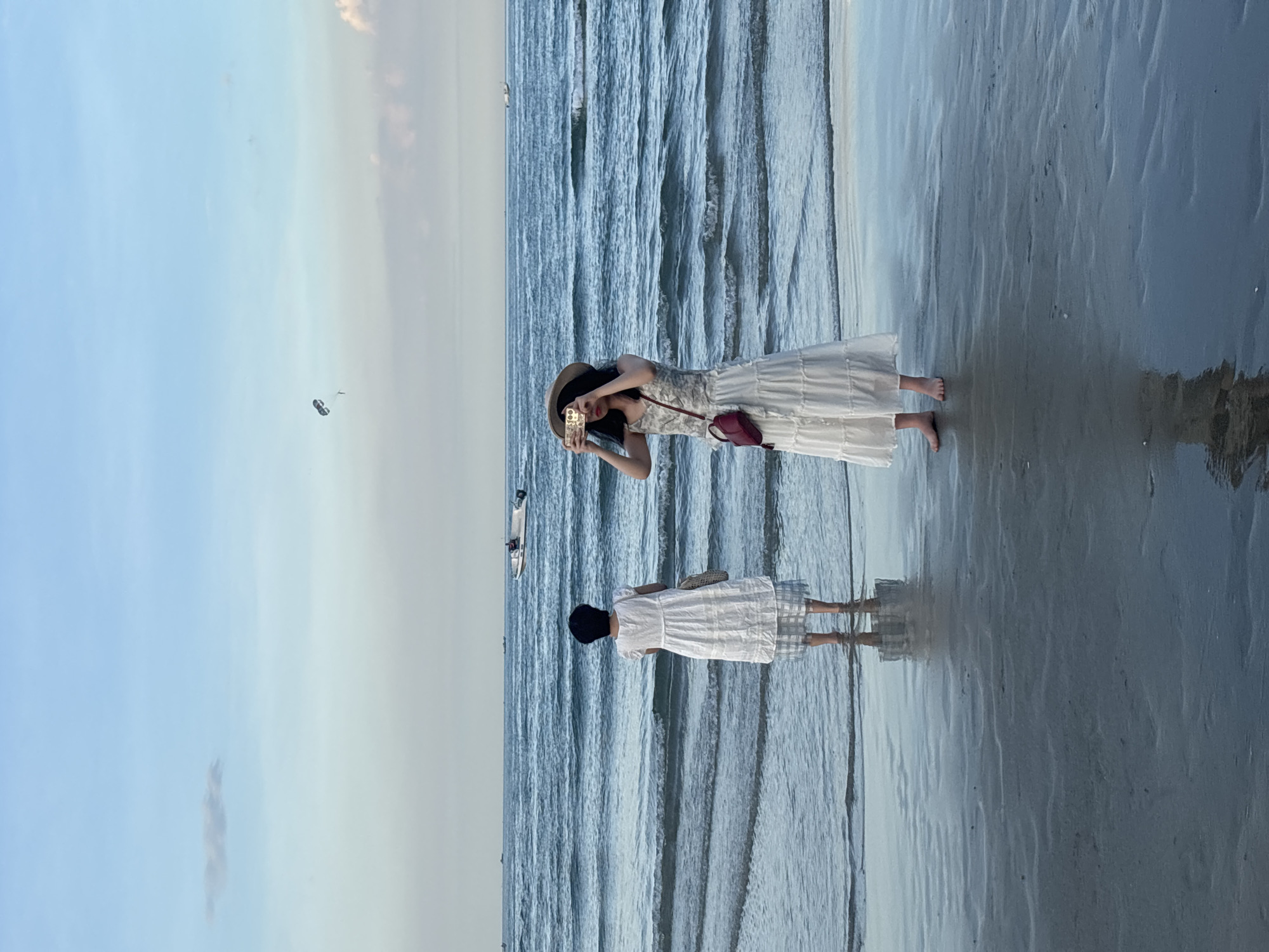This image depicts a serene beach scene at dusk in Đà Nẵng, Vietnam. Two women are visible standing in shallow water, facing the ocean. The woman on the right, wearing a white tiered dress and a straw hat, is holding up a mobile phone to take a picture. She has a small red bag slung across her shoulder. The woman on the left, also in a white dress, stands further back and faces away from the camera, observing the sea. The ocean is calm with gentle waves, reflecting the muted colors of the sky. A parasail is visible in the distance, suspended against the hazy blue sky. Several small boats are also scattered across the horizon. The beach itself is wet and reflective, creating mirror images of the figures standing in the water. The overall atmosphere is peaceful and tranquil, characteristic of a beach at the end of the day.