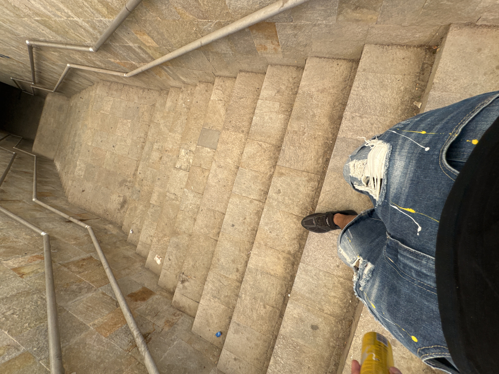 The image shows a first-person perspective looking down a set of outdoor stone stairs. Two metal handrails run along the sides of the stairs, angled slightly inward. The stairs are constructed from large, light-colored stone slabs with visible grout lines and some staining. There is some minor debris scattered on the steps, including a small blue plastic object on one step and some crumpled paper on another.
In the foreground, a person's legs are visible, wearing distressed blue denim jeans with rips and paint splatters of yellow and white. The person is wearing a dark-colored shoe on their right foot. They are holding a bright yellow cylindrical object, possibly a can, in their left hand. The lighting suggests it is daytime, with shadows cast by the handrails and the surrounding environment. The location in the city of Sofia, Bulgaria, is not discernible from the visual information in the image.