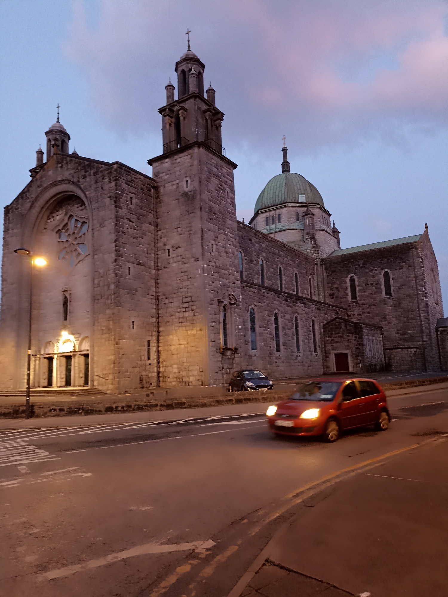 The image depicts the Galway Cathedral in the city of Galway, Ireland, at dusk. A red car is driving on the road in the foreground, its headlights illuminated. To the left of the road, a streetlamp is on. The cathedral is a large, stone building with a prominent dome and two bell towers. The sky is a muted blue with streaks of pink clouds. The scene is captured from a slightly low angle, looking up at the cathedral.