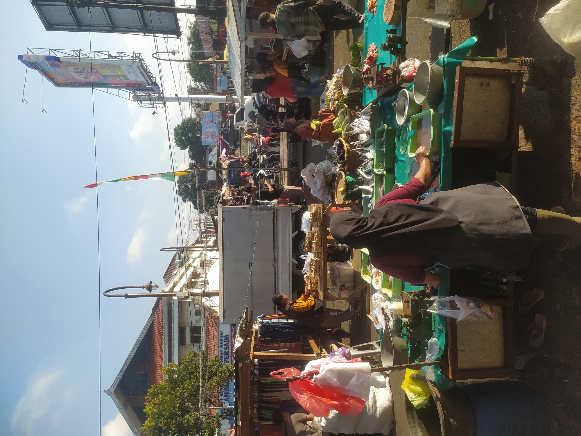 This is a medium shot of a bustling outdoor market, likely a fish market, on a sunny day. The scene is set in Kupang, Indonesia, as suggested by the context. In the foreground, a woman wearing a black hijab and an apron is attending to her stall. She is standing behind a green-covered table displaying various goods, including fish in green plastic containers, some raw meat, and various produce. A scale is visible on the table, indicating the transactional nature of the setting. Various plastic bags and bowls are scattered around her stall. Behind her, other vendors and customers are visible, engaged in their respective activities. To the left, a stall with hanging clothes is set up, with a woman sorting through them. Further back, a white truck is parked, and numerous other stalls and market structures are visible, extending into the background. The environment suggests a lively and active marketplace. The sky above is clear blue with a few wispy clouds, and a tall billboard advertising "TULASA" is prominently displayed on the right side of the image. Streetlights and power lines are also visible, adding to the urban market atmosphere.