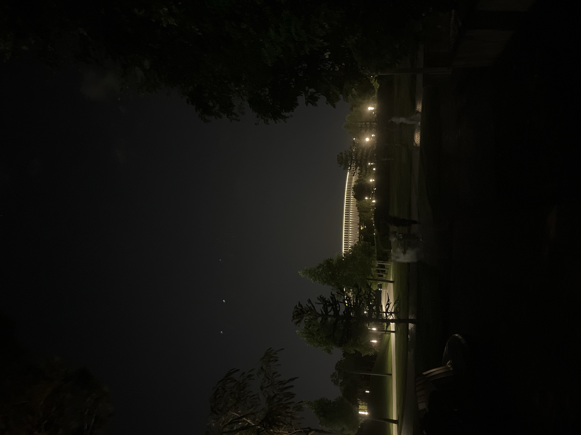 The image depicts a night view of the stadium "Krasnodar" in Krasnodar, Russia. The stadium, a prominent landmark, is visible in the background, illuminated by floodlights, with its distinctive architectural features clearly discernible. In the foreground, a park-like setting is present, with trees and pathways. Several lampposts are lit, casting pools of light on the ground and illuminating the foliage. There are a few indistinct figures of people walking in the park, their forms blurred due to the low light conditions and possibly motion. A park bench is visible in the lower left portion of the frame. The sky is dark, with a few faint lights that might be stars or distant aircraft. The overall atmosphere is serene and quiet, characteristic of a public space at night.