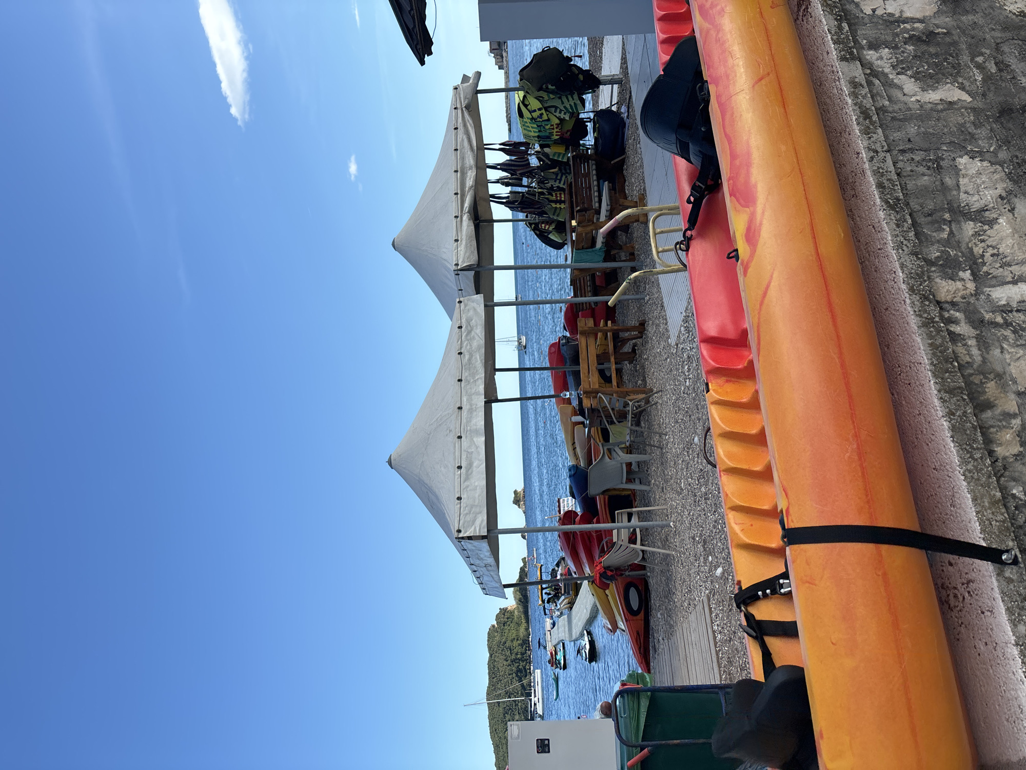 This image captures a vibrant waterfront scene in Budva, Montenegro, depicting a recreational area under a clear blue sky. The photograph is oriented vertically, with the horizon running down the left side.
In the immediate foreground on the right, several colorful kayaks are prominently displayed. These include bright red, orange, and yellow-orange kayaks, which appear to be stored or lined up along a rough concrete wall, secured with black straps. A black bag or backpack is visible on one of the kayaks.
Towards the center of the image, two large white canopy tents provide shade over a mixed surface of gravel and wooden decking. Beneath these canopies, tables and chairs are arranged, suggesting a seating or refreshment area. Notably, under the rightmost canopy, a rack holds multiple bright yellow-green life vests, indicating a facility for water sports.
Beyond the shaded area, the serene, deep blue sea stretches out, dotted with several boats, including sailboats and smaller vessels, anchored or navigating. Across the water, a lush, green, forested landmass, likely a part of the coastline or an island, adds to the scenic backdrop.
The sky is a brilliant, clear blue with only a few sparse, white wisps of clouds, suggesting excellent weather conditions. The bright daylight indicates it is likely morning or early afternoon. While no people are visible in the frame, the presence of kayaks, life vests, and seating areas strongly implies an active water sports rental or leisure spot. A white appliance with an unidentifiable text or logo is visible under the left canopy, possibly a cooler or refrigerator. The overall atmosphere is one of relaxation and recreational readiness, characteristic of a sunny coastal resort.