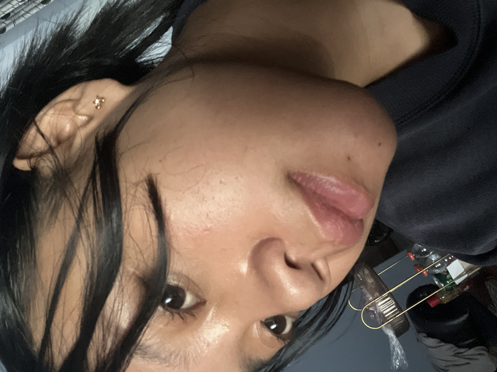 A close-up, angled shot features a young woman's face, capturing her left eye, nose, lips, and ear. Her dark hair is partially obscuring her face, with strands falling across her forehead. She is wearing a dark-colored sweatshirt and a small, gold stud earring in her left ear. The background is dimly lit and out of focus, showing what appears to be a shelf with various items, including bottles and plastic packaging. The lighting suggests an indoor setting. The image does not provide any information about the city of Jambearum, Indonesia, or any context related to that location.
