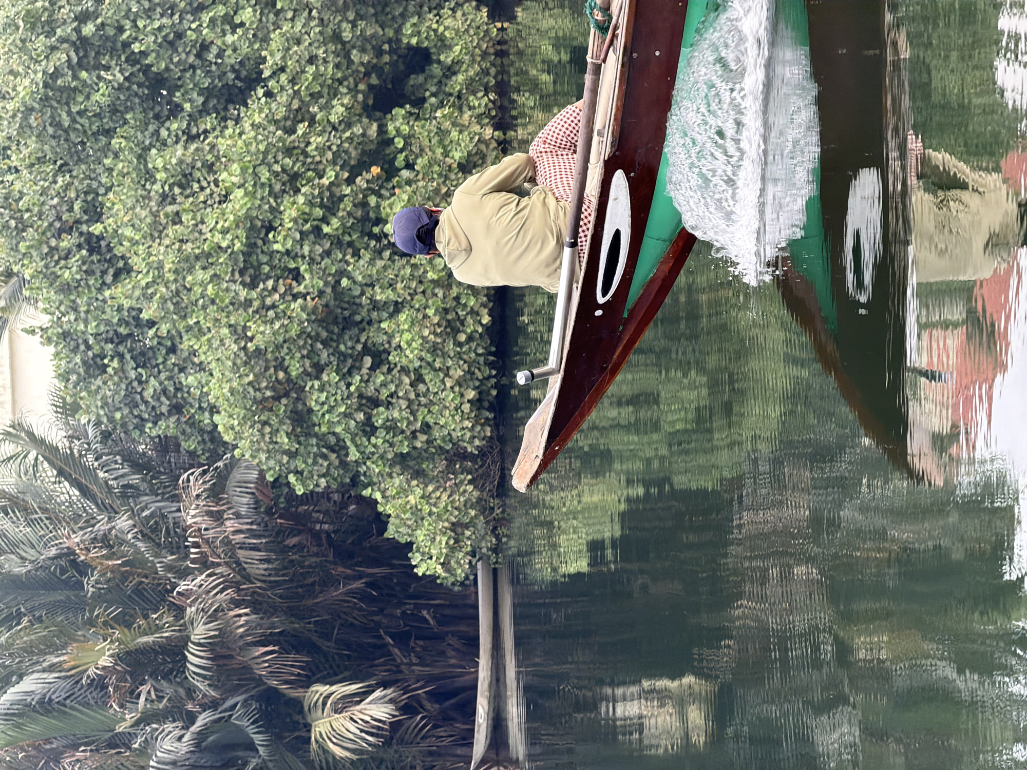 An outdoor, eye-level shot captures a person in a wooden boat navigating calm water, likely a river or canal, amidst lush tropical vegetation. The perspective suggests the viewer is also on the water, possibly in another boat.
The primary subject is a person, seen from behind, wearing a light olive or khaki-colored jacket, red and white checkered pants, and a dark blue baseball cap. They are seated in a traditional-looking narrow wooden boat, which has dark brown sides and a prominent green section along its upper edge. A white, elongated object is visible on the inner side of the boat. An oar or pole handle extends upwards near the person.
The boat is moving, creating a clear white wake on the water surface at its front. The water is dark and highly reflective, showing distorted but recognizable reflections of the boat, the person, and the surrounding foliage. The reflections are a significant visual element, mimicking the upper half of the scene upside down, but with a shimmering, painterly quality due to the water's movement.
The setting is characterized by dense, vibrant greenery. On the left side, a large mass of small-leaved green trees dominates, while closer to the water, darker, more textured palm fronds are visible. The overall impression is one of a serene, natural environment.
The time of day appears to be daytime, possibly under an overcast sky or in dappled sunlight, given the even lighting and lack of harsh shadows. There is no visible text in the image. The scene evokes a sense of tranquil travel through a verdant landscape, typical of waterways in regions like Hội An Ward, Vietnam.