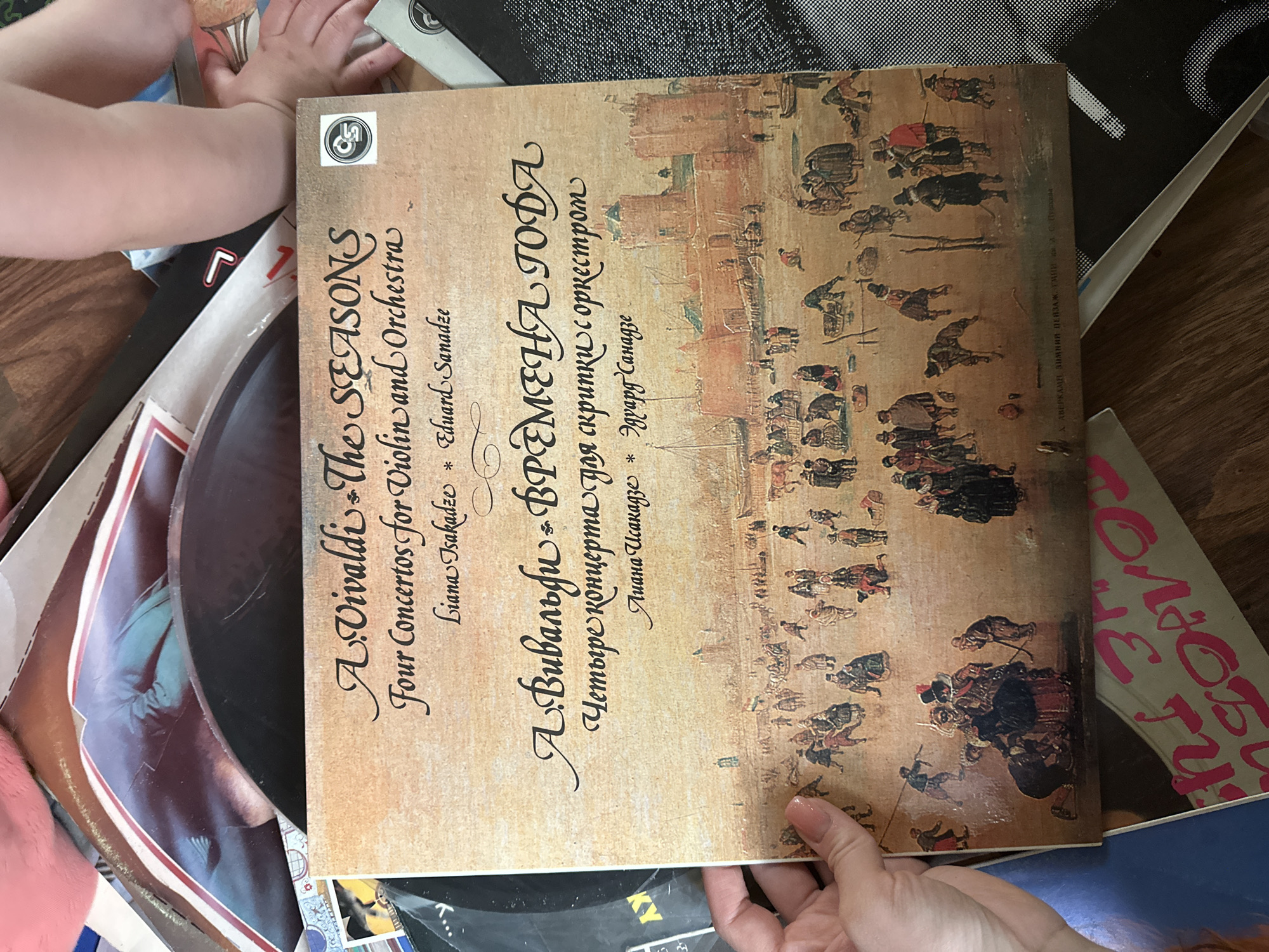 A high-angle shot shows a collection of vinyl records on a wooden floor. The focal point is a record sleeve displaying "A. Vivaldi - The Seasons", which translates to "ВРЕМЕНА ГОДА" in Russian. It is described as "Four Concertos for Violin and Orchestra", with names Liana Tsakadze and Eduard Sanadze. The sleeve's artwork features a period scene of people walking outside buildings. The artwork is “Avercamps Winter Landscape”. Hands are visible holding the record sleeve and other records in the stack. It is impossible to determine the location in Nizhny Novgorod, Russia, without additional context.