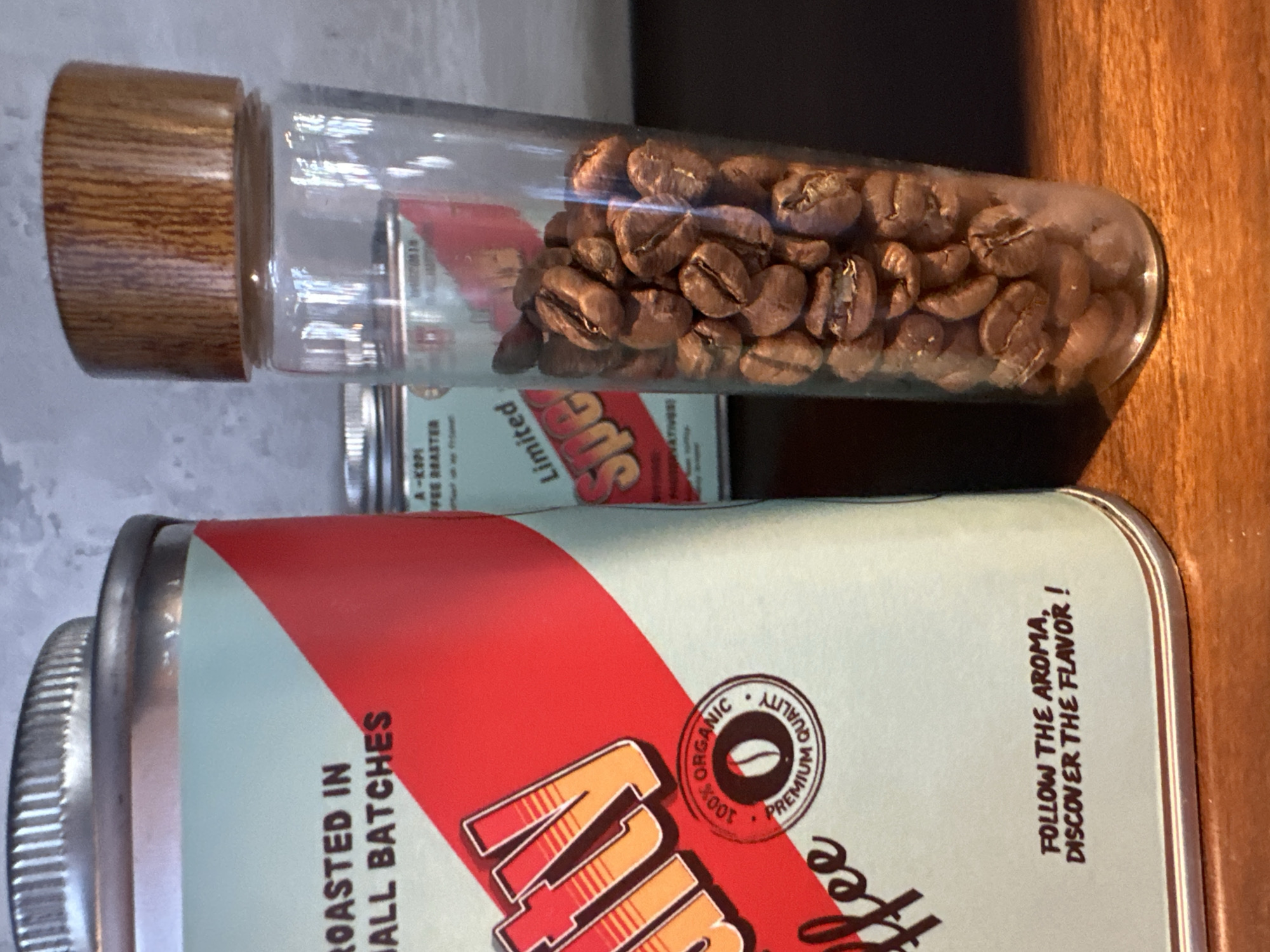 A close-up shot shows a large tin of coffee and a glass container filled with coffee beans. The tin, with a light blue and red design, has text that reads "ROASTED IN SMALL BATCHES" and "COFFEE". A circular emblem on the tin says "100% ORGANIC PREMIUM QUALITY". The glass container, topped with a wooden lid, displays a portion of coffee beans. In the background, another smaller tin is partially visible. The scene is set on a wooden surface with a textured gray wall behind. No specific location in Da Nang, Vietnam, is identifiable from the image.