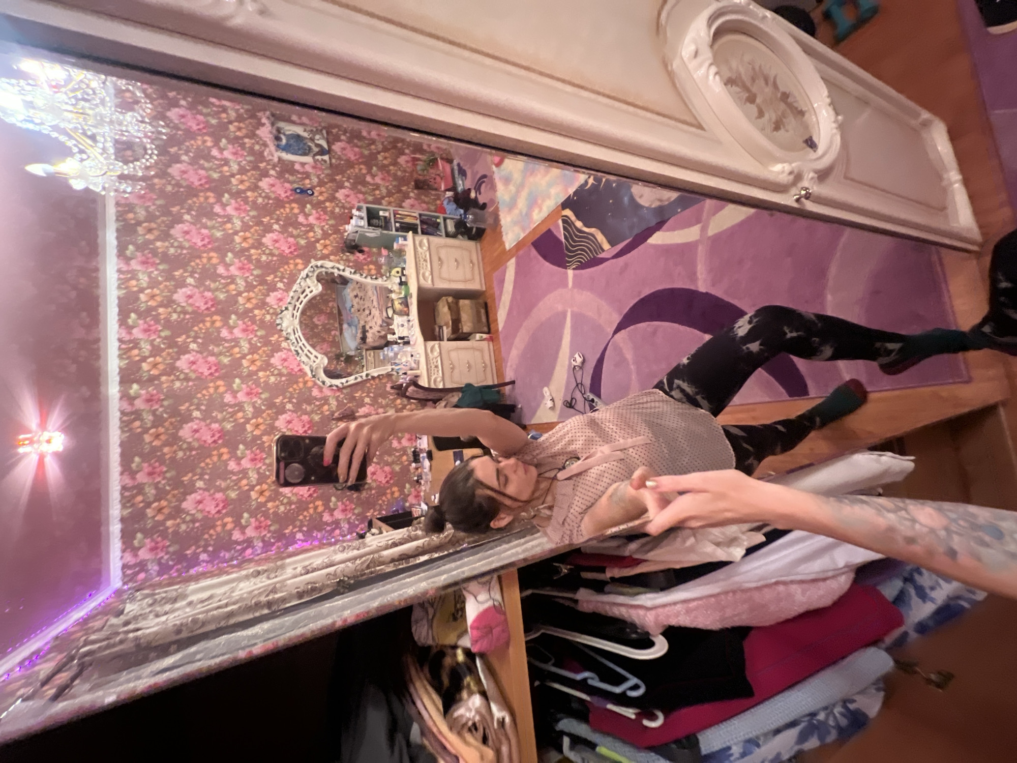 In a bedroom with floral wallpaper and pink LED strip lighting, a woman in a pink polka-dot shirt and patterned leggings stands in front of a full-length mirror, taking a selfie with her smartphone. The room is cluttered with clothes on hangers to the left of the mirror, and a vanity with a heart-shaped mirror, various items, and a small bookshelf are visible in the reflection. A purple rug with circular patterns covers part of the wooden floor. The overall impression is of a personal and perhaps bohemian living space in the city of Krasnoe-na-Volge, Russia.