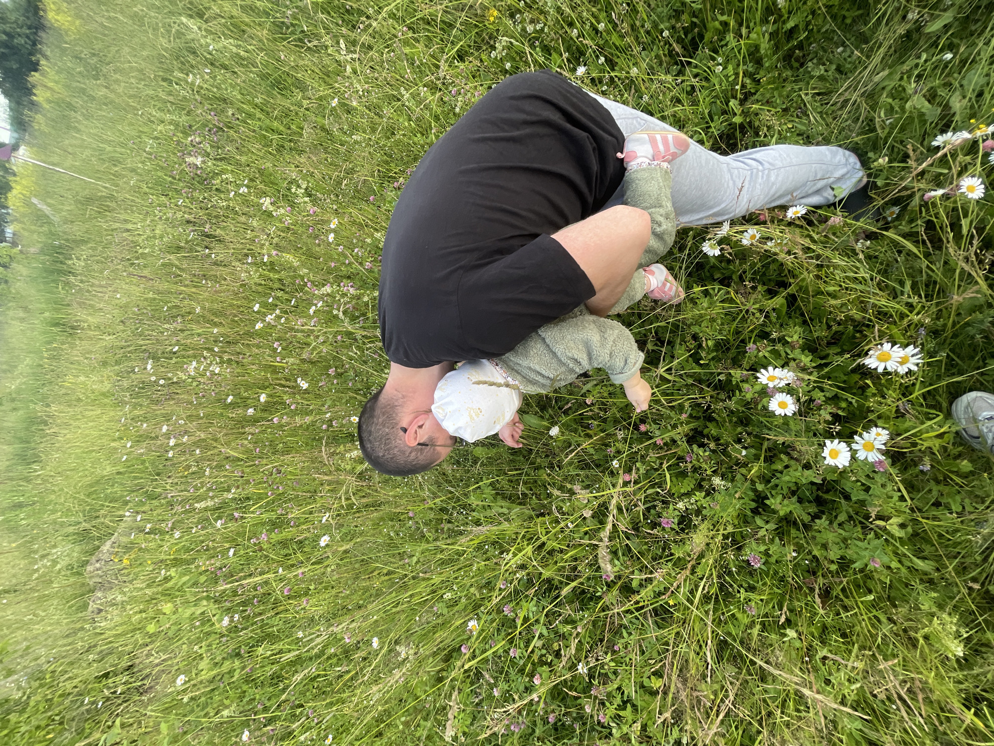 A man in a black t-shirt and grey sweatpants is holding a baby in his arms, surrounded by tall green grass and wildflowers. The baby is wearing a white hat and a light green textured outfit, with pink and white shoes. They are in a grassy field with numerous small white and pink flowers, some of which are daisies. The man appears to be looking down at the baby and the grass. The scene is outdoors in what appears to be a natural, unmanicured area, possibly a field or meadow on the outskirts of a settlement. The location is specified as the city of Podshivalovo, Russia.