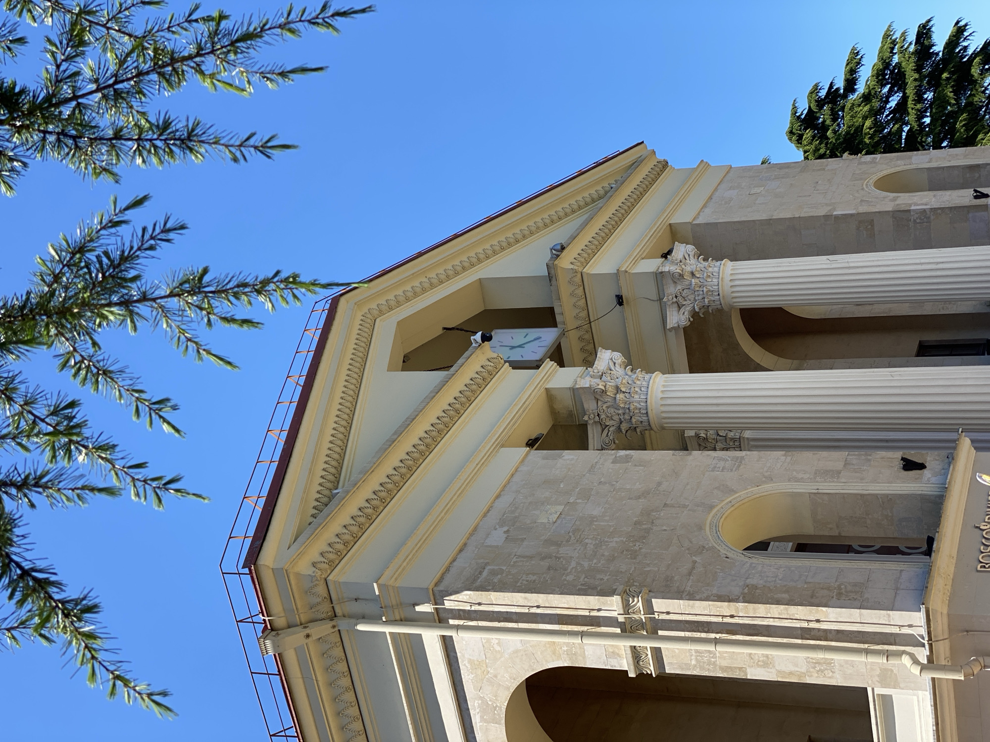This media file captures a low-angle view of a grand building's upper facade set against a clear blue sky. In the foreground on the left, several branches of a conifer tree, possibly a cedar or fir, extend into the frame, their green needles contrasting with the vibrant sky.
The building exhibits classical architecture, featuring prominent fluted columns with ornate, possibly Corinthian, capitals. A pediment-like structure crowns the facade, adorned with decorative friezes and molding. Beneath the eaves of this structure, a square white clock with black hands is clearly visible. The walls are constructed from light-colored stone blocks, showing signs of weathering. Arched openings and a metal gutter pipe are visible towards the lower right. A red-brown metal safety railing runs along the roofline on the left.
The bright daylight indicates a clear, sunny day, with strong shadows defining the architectural details. Part of a sign on the building's lower right edge shows the partial text "BOSCOV," likely belonging to "BOSCO di Ciliegi," a luxury brand often associated with retail or resorts. The setting is an elegant outdoor environment in Sochi, Russia.