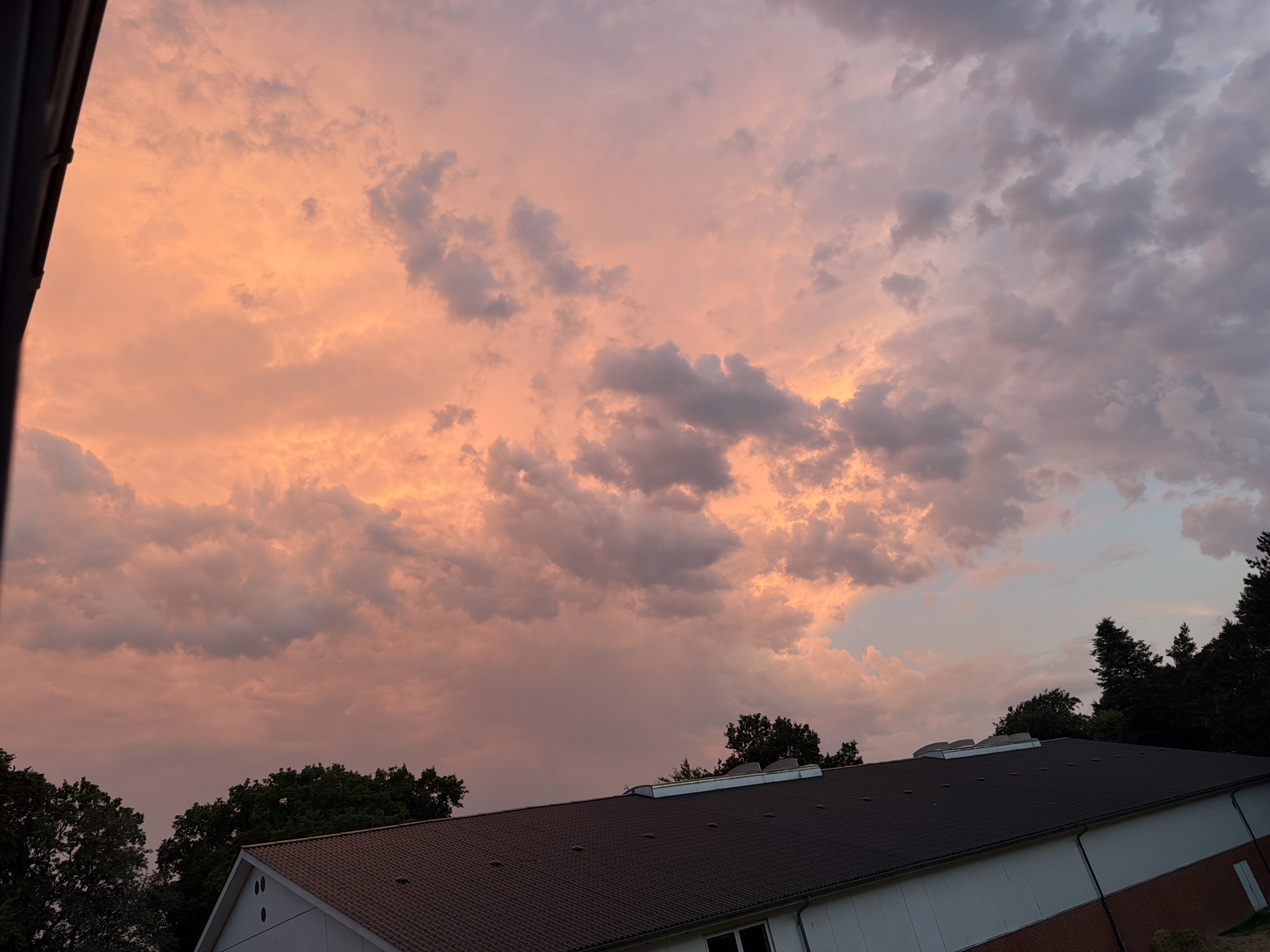 A dramatic sunset paints the sky in vibrant hues of orange and pink, casting a warm glow over the landscape of Esbønderup, Denmark. Clouds, illuminated by the setting sun, are a mixture of soft pinks, oranges, and darker grays. In the foreground, the dark, angled roof of a building dominates the lower portion of the frame. The building appears to be a large, modern structure with white siding and a visible brick foundation. A line of dark green trees forms a silhouette against the colorful sky, bordering the building on the left and extending across the middle ground to the right. The overall scene is one of natural beauty and serene twilight in a Danish town.