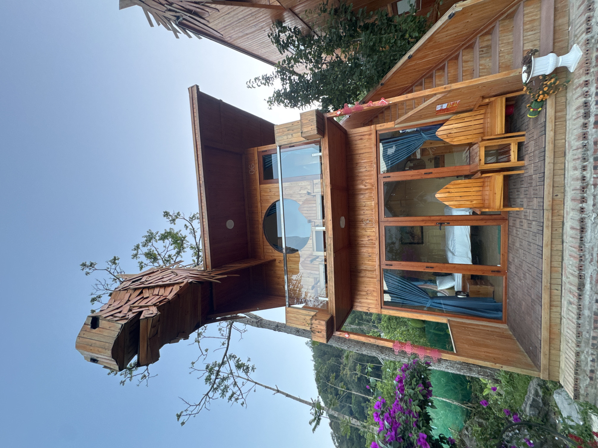 The image shows an unusual wooden structure in Bắc Hà, Vietnam, likely a hotel room or cabin. The structure is two stories tall, with a unique exterior design that includes a rounded window and an exterior that resembles a stylized animal head with shingled "scales." The cabin sits elevated, connected to another building with wooden steps, indicating it's part of a larger complex. Outside the cabin, there is lush greenery, with purple flowers growing near the base. There are trees that seem to grow through the lower level of the building. A glimpse of a bed inside the cabin is visible through the window. The sky is overcast.