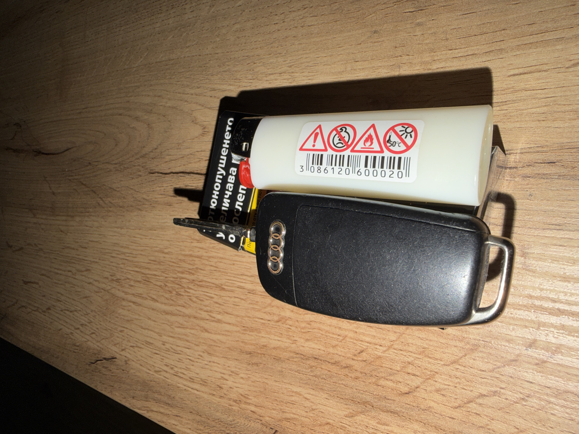 A close-up, top-down shot shows an Audi car key and a white disposable lighter lying on a wooden surface. The Audi key is black with a silver Audi logo. The lighter has several warning symbols, including a flame and a temperature warning. Behind the key and lighter is a black and yellow package, likely for tobacco products, with text in Bulgarian. The wooden surface appears to be a table or desk. The lighting casts shadows to the right of the objects, suggesting a light source from the left. The overall scene is static and appears to be indoors. There is no indication of the specific location within Plovdiv, Bulgaria.