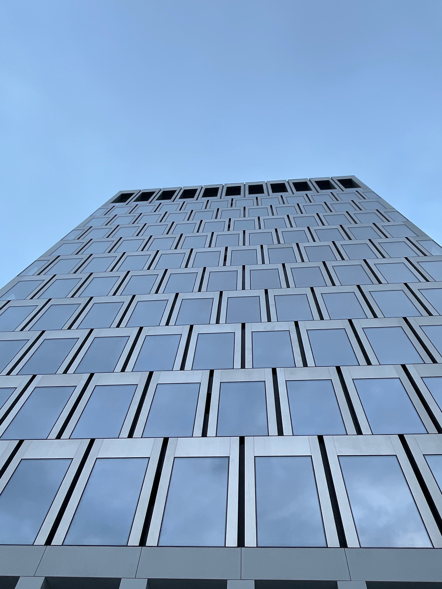 This is a low-angle, full-frame photograph of a modern skyscraper in Moscow, Russia. The building's facade is composed of numerous rectangular windows with dark frames, arranged in a repeating pattern that ascends upwards and to the left. The windows reflect a cloudy, pale blue sky. The perspective emphasizes the building's height and geometric design, with the top of the structure appearing to reach into the vast expanse of the sky. There are no people or other distinguishing features visible in the image.
