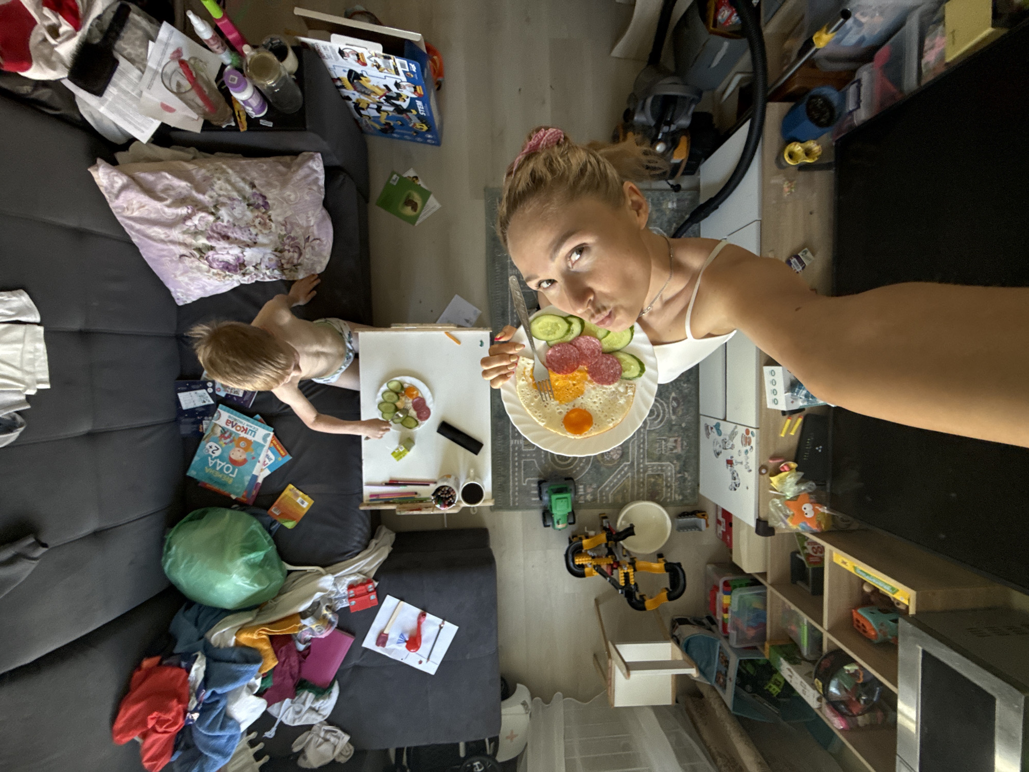 A woman is taking a selfie indoors with a plate of food in her hand. The plate contains an egg, cucumbers, and salami. A child is visible to the left of the woman, also with a plate of food. The scene is cluttered with various items scattered around the living room, including toys, books, clothes, a vacuum cleaner, and furniture. There is also a TV stand with various items in the shelves. The location is in an apartment in the city of Chaikovsky, Russia.