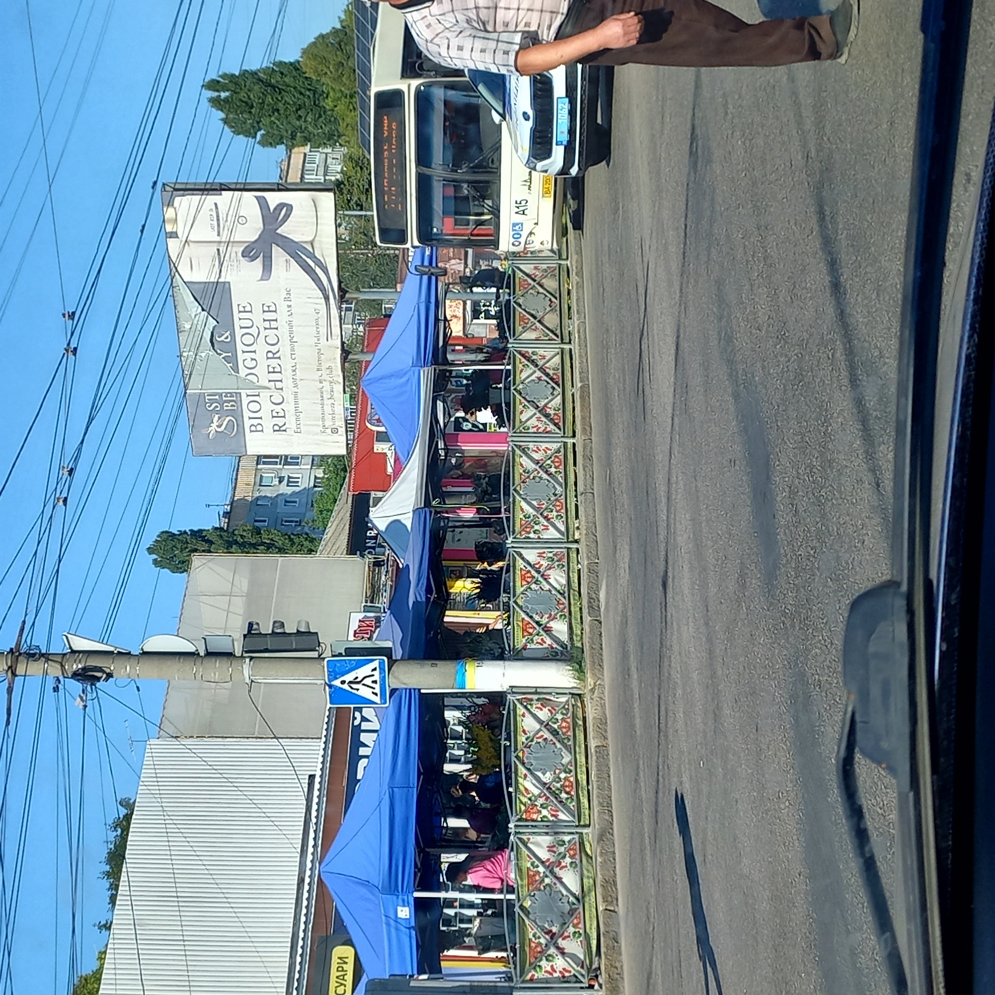 This image depicts a street scene in Kropyvnytskyi, Ukraine. In the foreground, the dashboard and windshield of a vehicle are visible, suggesting the photo was taken from inside a car. The street surface is asphalt. To the right, a man wearing a plaid shirt and brown pants is walking across the road. Beyond the road, there are several tents set up, likely for market stalls, with people inside. A pedestrian crossing sign is visible on a utility pole. Further in the background, a bus is stopped at what appears to be a bus stop, and a white car is parked nearby. A large billboard with text and an image of a bow is also visible in the distance. The sky is blue with visible overhead power lines.