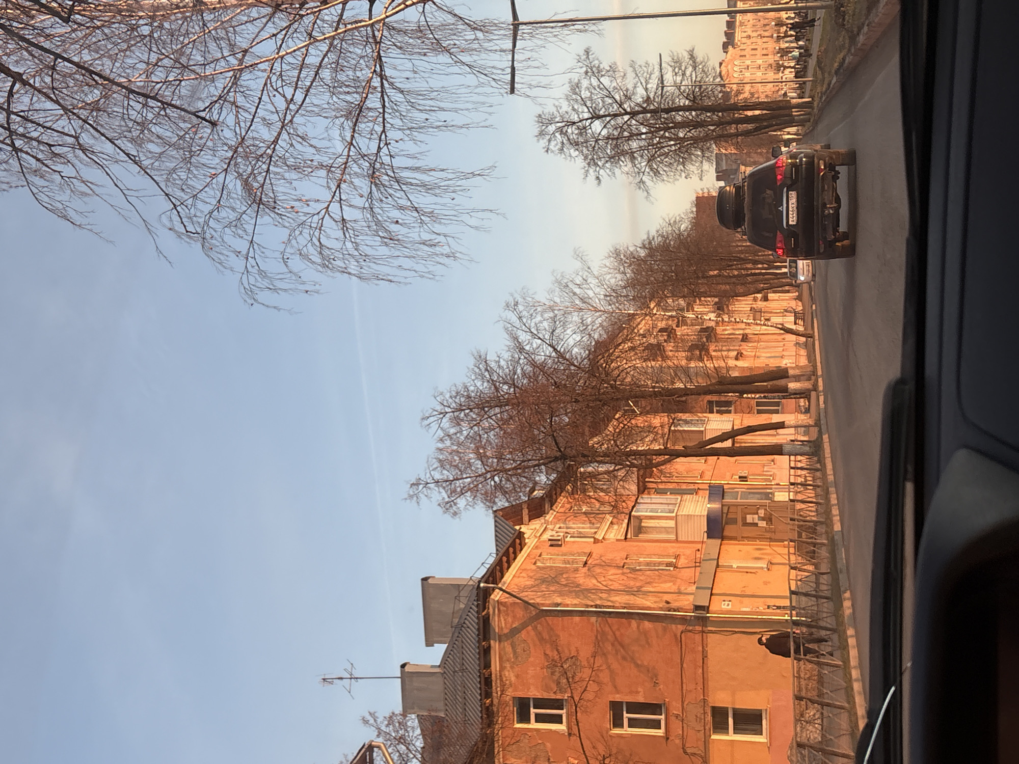 The image captures a street scene in what appears to be late autumn or early spring, given the bare deciduous trees. The perspective is from within a vehicle, looking forward and slightly to the right, with part of the vehicle's interior visible on the far right.
A multi-story street runs into the distance, lined on the right by buildings illuminated by warm, low-angle sunlight, indicating either early morning or late afternoon. The buildings have an orange or peach-colored facade, with visible windows and some balconies. Antennas are visible on the rooftops.
In the foreground and middle ground of the street is a dark-colored SUV, possibly a Mitsubishi, with a spare tire mounted on its roof rack. The vehicle's rear license plate, "A446KT 182", is clearly visible, with "182" being the Russian region code for the Udmurt Republic, confirming the location as Votkinsk, Russia.
Bare-branched trees line both sides of the road, casting long shadows. The sky is clear and light blue, with a slight haze towards the horizon. Faint figures of people can be seen walking on the distant sidewalk next to the buildings. A lone dark figure is also visible on a building's balcony or roof ledge on the right side of the image.