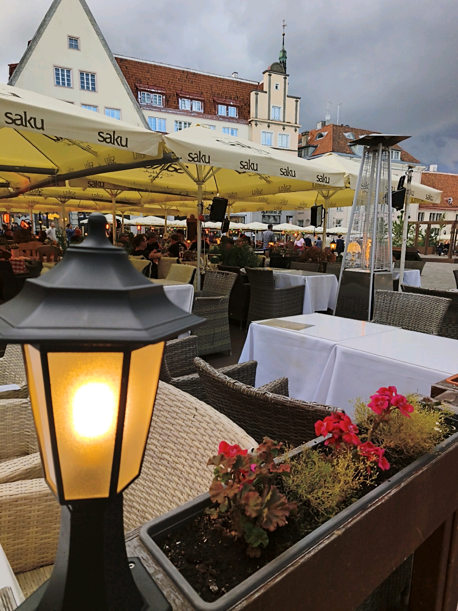 The image shows an outdoor seating area of a restaurant or cafe in Tallinn, Estonia, likely in the Old Town. In the foreground, a black lamppost with an illuminated yellow glass shade is visible. Behind it, wicker chairs and tables covered with white tablecloths are arranged. Yellow umbrellas with the "Saku" logo are spread overhead, providing shade. In the background, traditional European buildings with red-tiled roofs and light-colored facades are visible. Several people are seated at tables in the distance, engaged in dining or conversation. A tall, pyramid-shaped outdoor heater is also present in the seating area. The sky is overcast, suggesting a cloudy day.