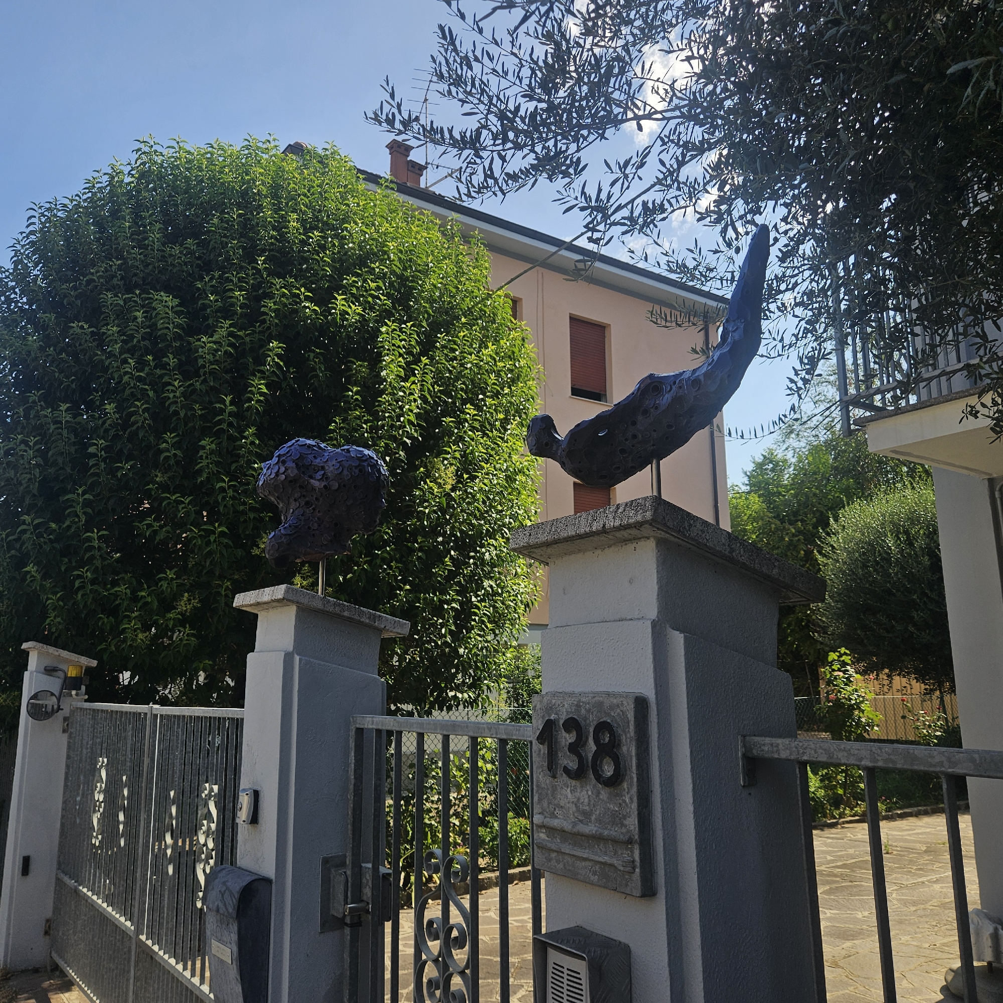 This is a medium shot, taken outdoors on a sunny day, showing an entrance gate and fence in Modena, Italy. The gate is made of metal with decorative scrollwork. To the left of the gate, on a concrete pillar, is a dark-colored, abstract sculpture resembling a cluster of bubbles. To the right of the gate, also on a concrete pillar, is another dark-colored, abstract sculpture that appears to be a stylized human figure in a diving or falling pose. This sculpture has visible holes and textured surfaces. A gray concrete pillar to the right of the figure sculpture has the number "138" mounted on it. Behind the gate and sculptures, a large, dense green tree is visible, and beyond that, a light pink building with red-shuttered windows. An olive tree with dark leaves partially obscures the top of the image. The ground in front of the gate is paved with stone tiles.