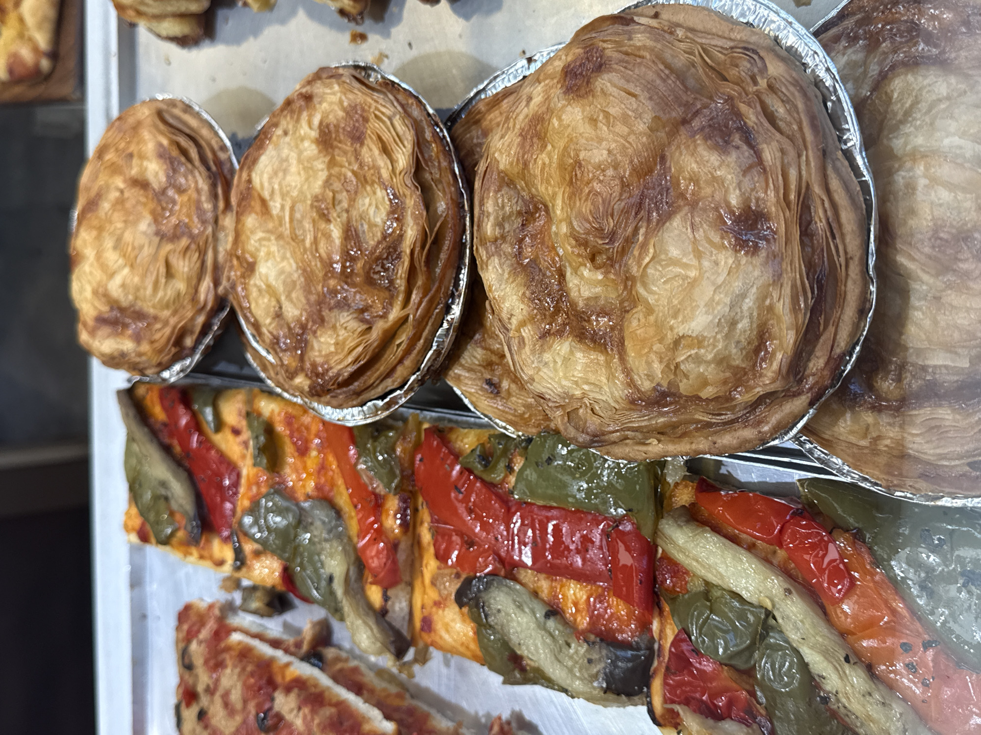 The image presents a detailed view of various baked goods displayed on a reflective, silver-colored metallic tray, indicative of a commercial food environment such as a bakery or deli in Unknown, Unknown. On the left side of the tray, several rectangular slices of focaccia or a similar flatbread are arranged. These slices are topped with large pieces of roasted vegetables, including vibrant red bell peppers, green bell peppers, and elongated, pale green-grey strips resembling roasted eggplant. The vegetable toppings appear glistening, possibly from oil or a glaze. A partially visible slice of pizza, featuring red sauce and dark circular toppings, is located in the upper left quadrant. On the right side of the tray, a stack of at least five individual, round pastries is prominently featured. Each pastry rests in a circular aluminum foil cup and exhibits a golden-brown, highly layered, and flaky crust, consistent with puff pastry. The crusts show varying degrees of browning and a slightly glossy finish. The background is out of focus, suggesting the interior of a display case or counter. No individuals or specific actions are depicted within the scene.
