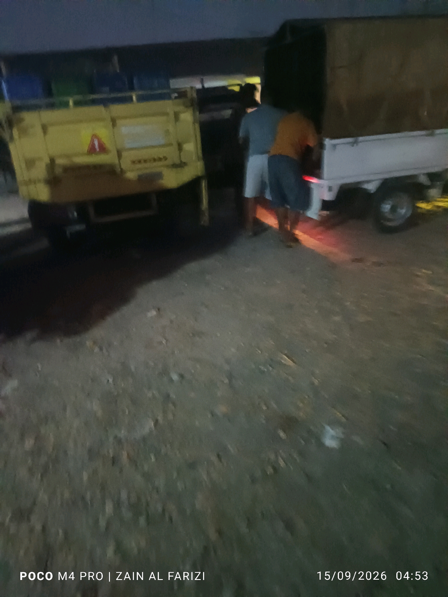 In the city of Arosbaya, Indonesia, a yellow truck is parked on a dirt road, its bed filled with various items, possibly goods or debris. A white truck with a canvas cover is parked to the right of the yellow truck. Three individuals are visible near the white truck; one in a gray shirt and white shorts, another in an orange shirt and blue shorts, and a third person whose attire is less clear due to motion blur. They appear to be engaged in an activity related to the trucks, though the specific nature of their actions is not discernible. The environment suggests it is either early morning or late evening, as indicated by the dim lighting. The overall scene depicts a functional, possibly logistical, activity occurring on a utilitarian roadway.