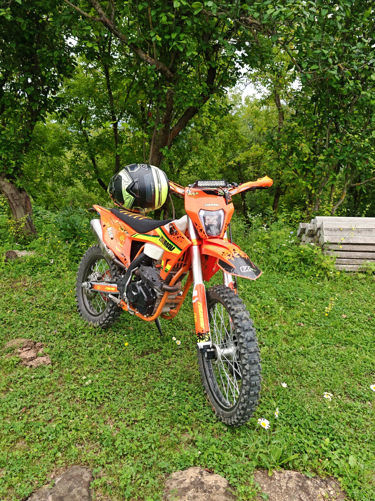 An orange dirt bike is parked on a grassy knoll in a wooded area. A black and yellow helmet rests on the handlebars. In the background, a stack of concrete slabs is visible. The scene is set in the city of Sherdy-Mokhk, Russia.