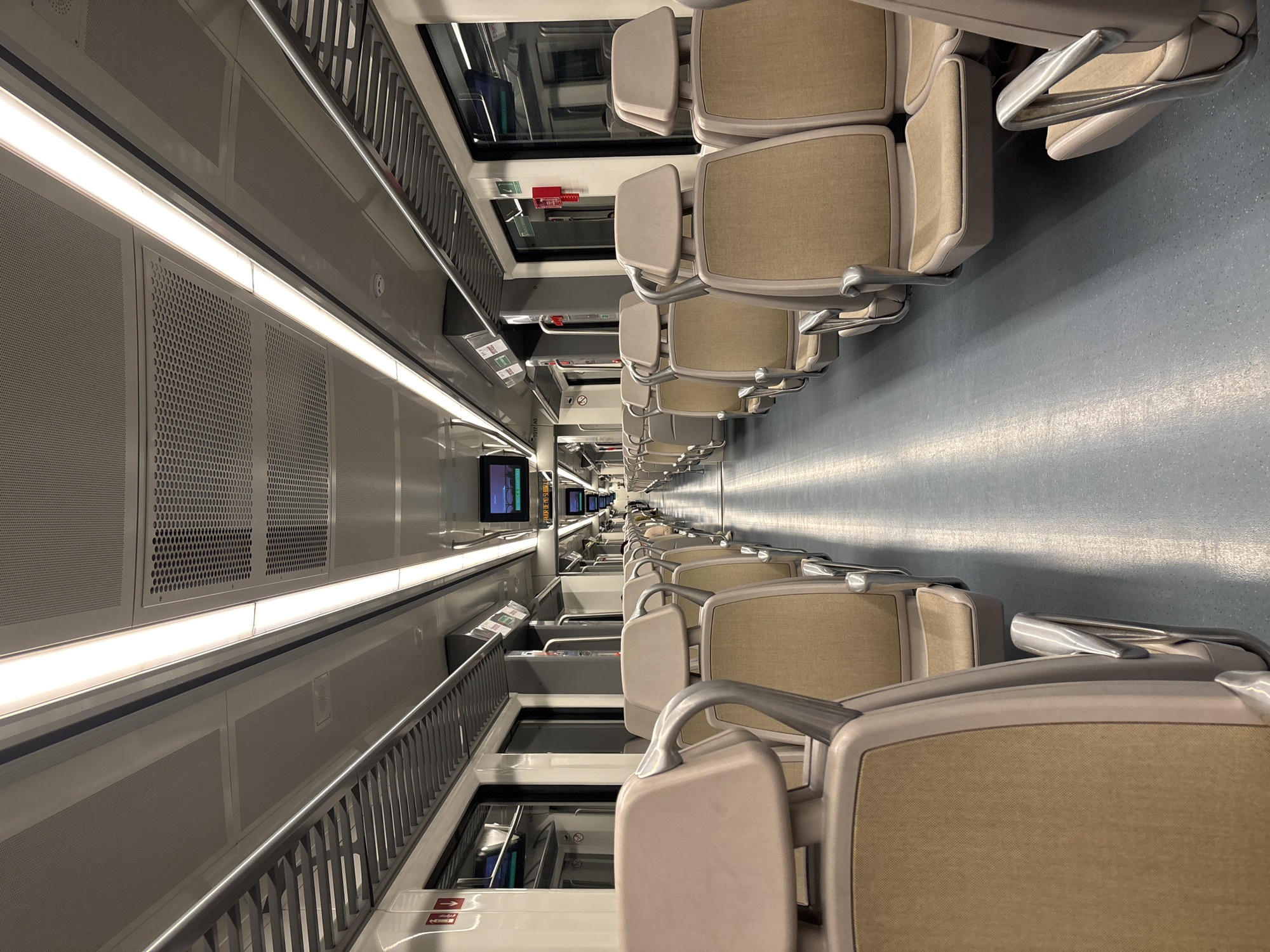 The image shows the interior of a train car, likely a commuter or subway train. Rows of empty seats upholstered in a light brown or beige fabric are visible on either side of a central aisle, receding into the distance. The seats have light gray or beige frames and armrests. The floor is a light blue-gray. The walls of the train car are light gray and feature perforated panels and horizontal lighting fixtures. Windows are present above the seats, with safety and information signage visible near them. Several video screens are embedded in the upper portion of the train car, displaying information. The composition is taken from a perspective at one end of the train car, looking down the length of the aisle. It is not possible to determine whether this is the city of La Marquesa, Mexico based on the provided image.