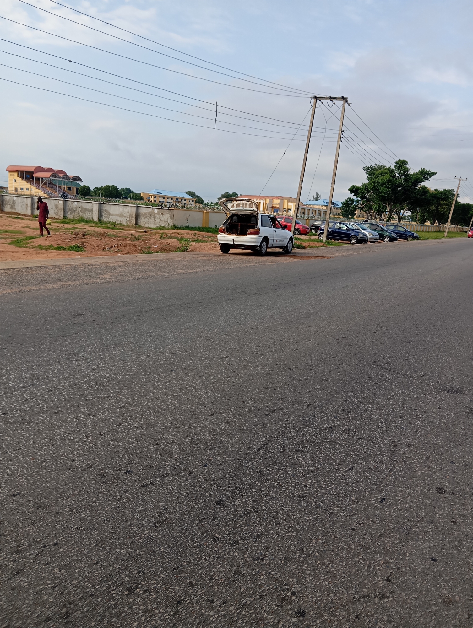 A white hatchback car with its trunk open is parked on the side of a paved road in Jalingo, Nigeria. Beside the car, a person wearing a maroon traditional outfit is walking on a dirt path towards a concrete wall topped with barbed wire. Behind the wall, several other cars are parked in a row, and further back are buildings, including a large structure with a red roof and tiered seating, resembling a stadium or conference center, and another building with a blue roof. Utility poles with numerous wires are visible overhead, and the sky is partly cloudy. The road in the foreground is dark asphalt with visible gravel.