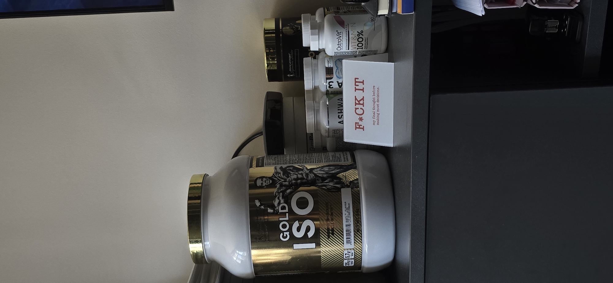 The image shows a close-up, eye-level shot of a shelf holding various health and fitness products, along with a humorous sign. The items are arranged on a dark gray surface, likely a shelf in a home or gym environment. The background is a plain, light-colored wall.
Dominating the left side of the image is a large, white and gold tub of "GOLD ISO" protein powder. The label features a muscular silhouette of a bodybuilder. To the right of the protein tub are several smaller bottles of supplements. One prominent bottle is from "OstroVit" and contains "VIT & MIN 100%".
In front of the supplement bottles, a small white card is placed. The card has a red, bold text that reads "F*CK IT" and below it, in smaller red text, "my final thought before making most decisions."
The scene suggests a personal space where fitness and perhaps a sense of humor are valued. There is no indication of the city of Vratsa, Bulgaria, or any specific location details in the image. The lighting appears to be natural, coming from the front and slightly to the left, casting soft shadows. The image is in focus and captures the details of the products and the sign clearly.